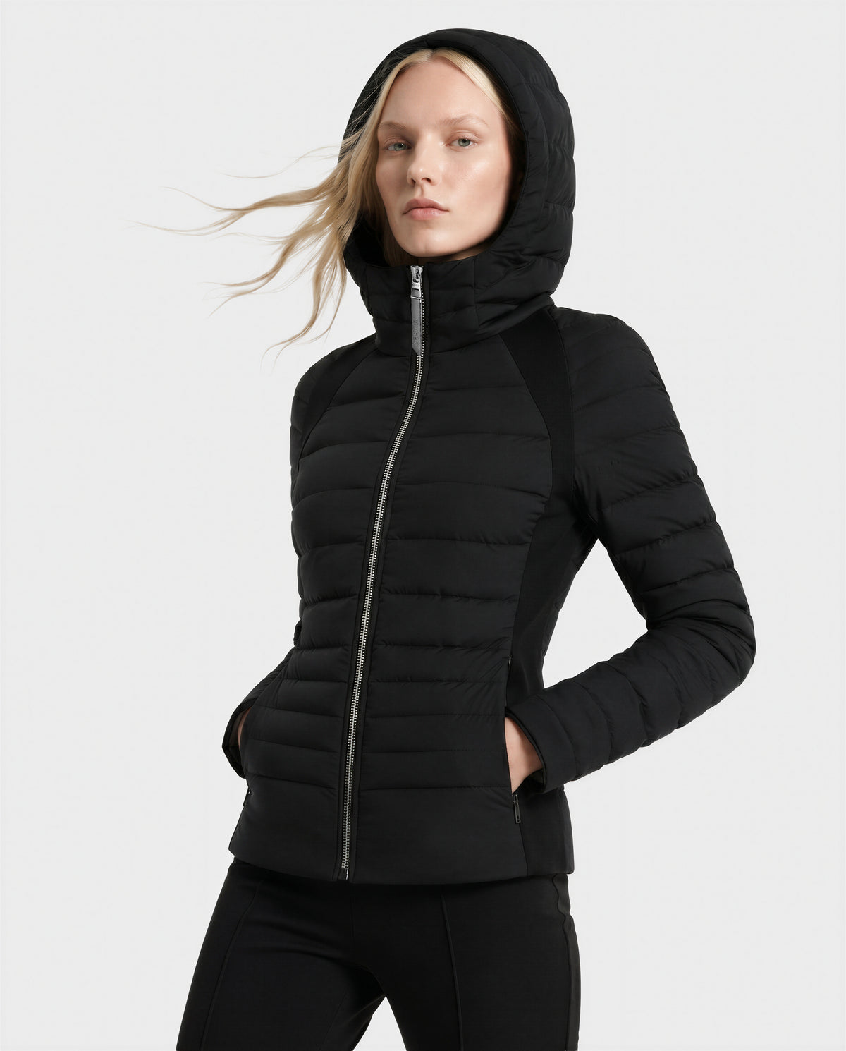 A person with long blonde hair wears the Rudsak LENA - L124005 Black quilted jacket and black pants, standing with hands in pockets against a plain light background.