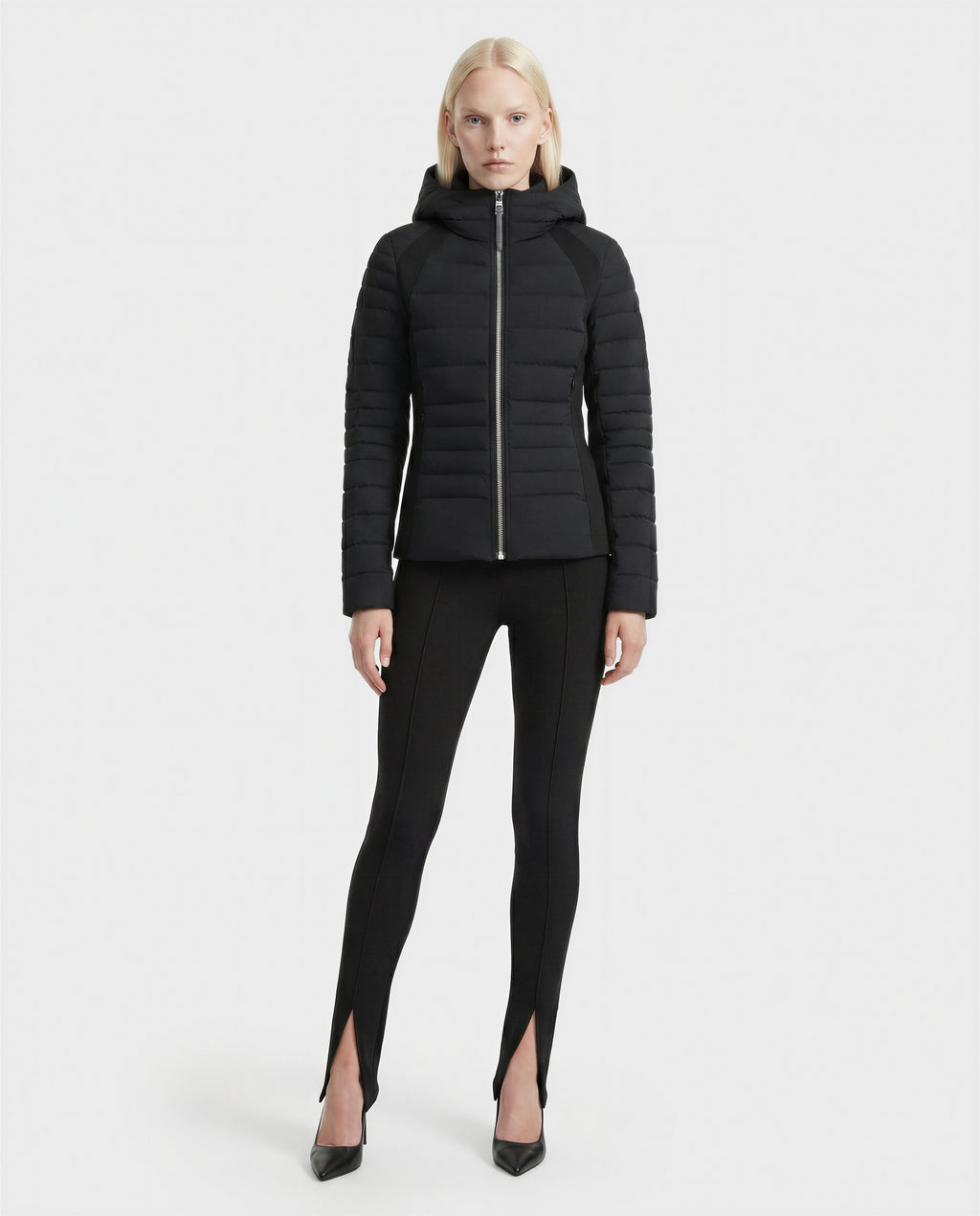 A person with straight blonde hair wears the Rudsak LENA - L124005 Black fitted quilted jacket, paired with black leggings featuring front ankle slits and black pointed-toe heels, standing against a plain white background.