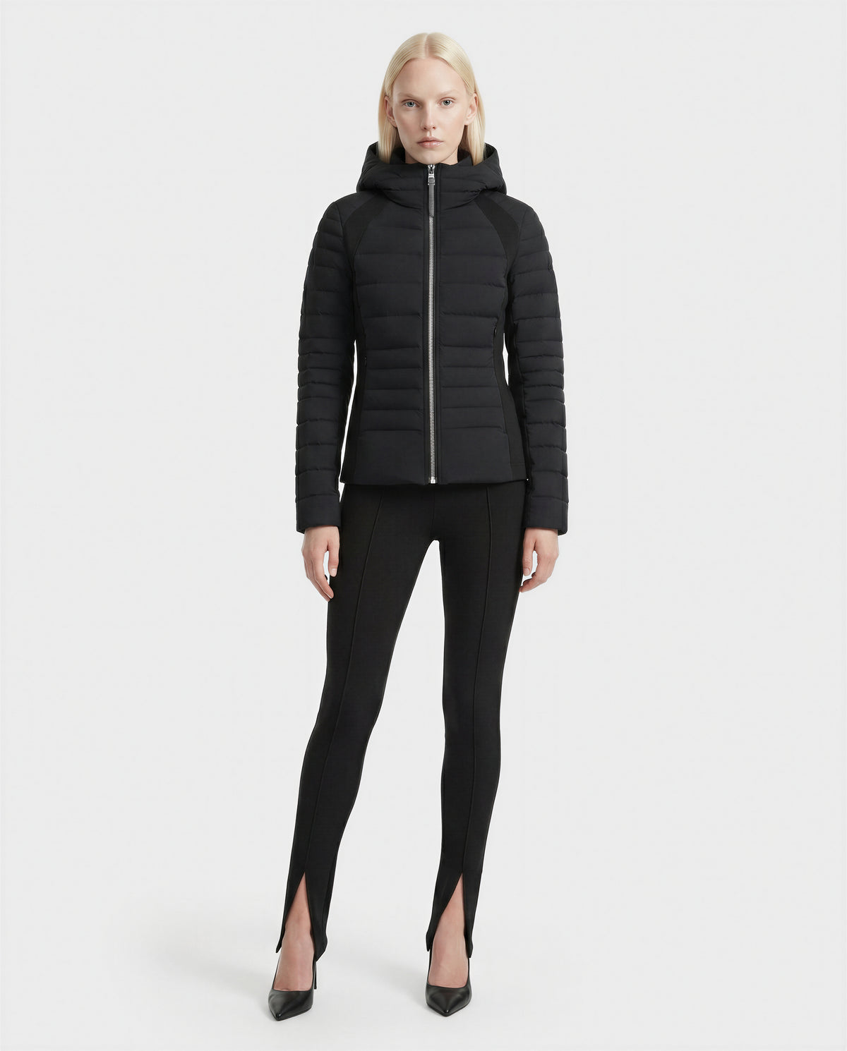 A person with straight blonde hair wears the Rudsak LENA - L124005 Black fitted quilted jacket, paired with black leggings featuring front ankle slits and black pointed-toe heels, standing against a plain white background.