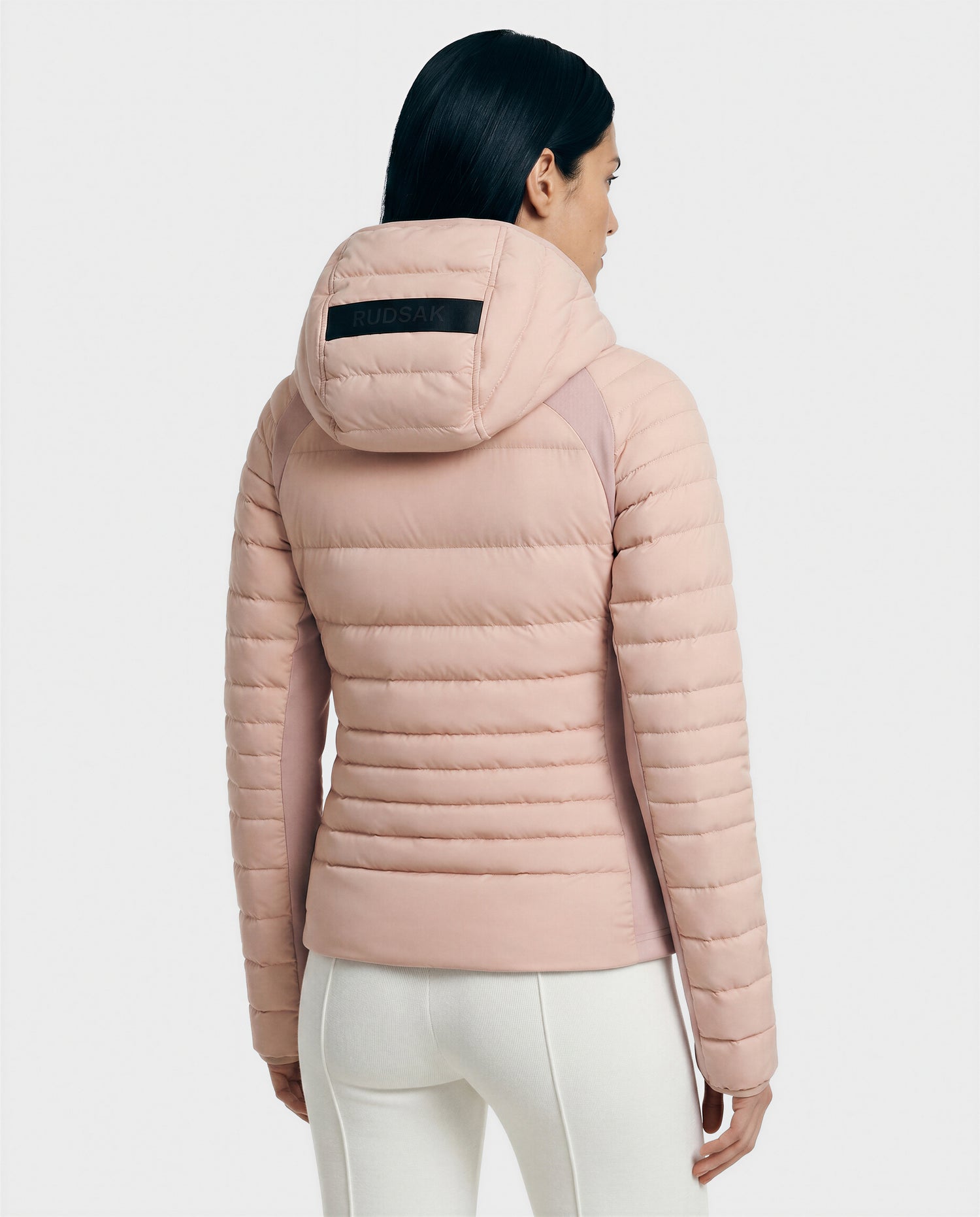 A woman with long dark hair is seen from behind, wearing the Rudsak LENA - L124005 Soft Pink women's down jacket with a label on the hood and paired with white fitted pants, standing against a plain light background.