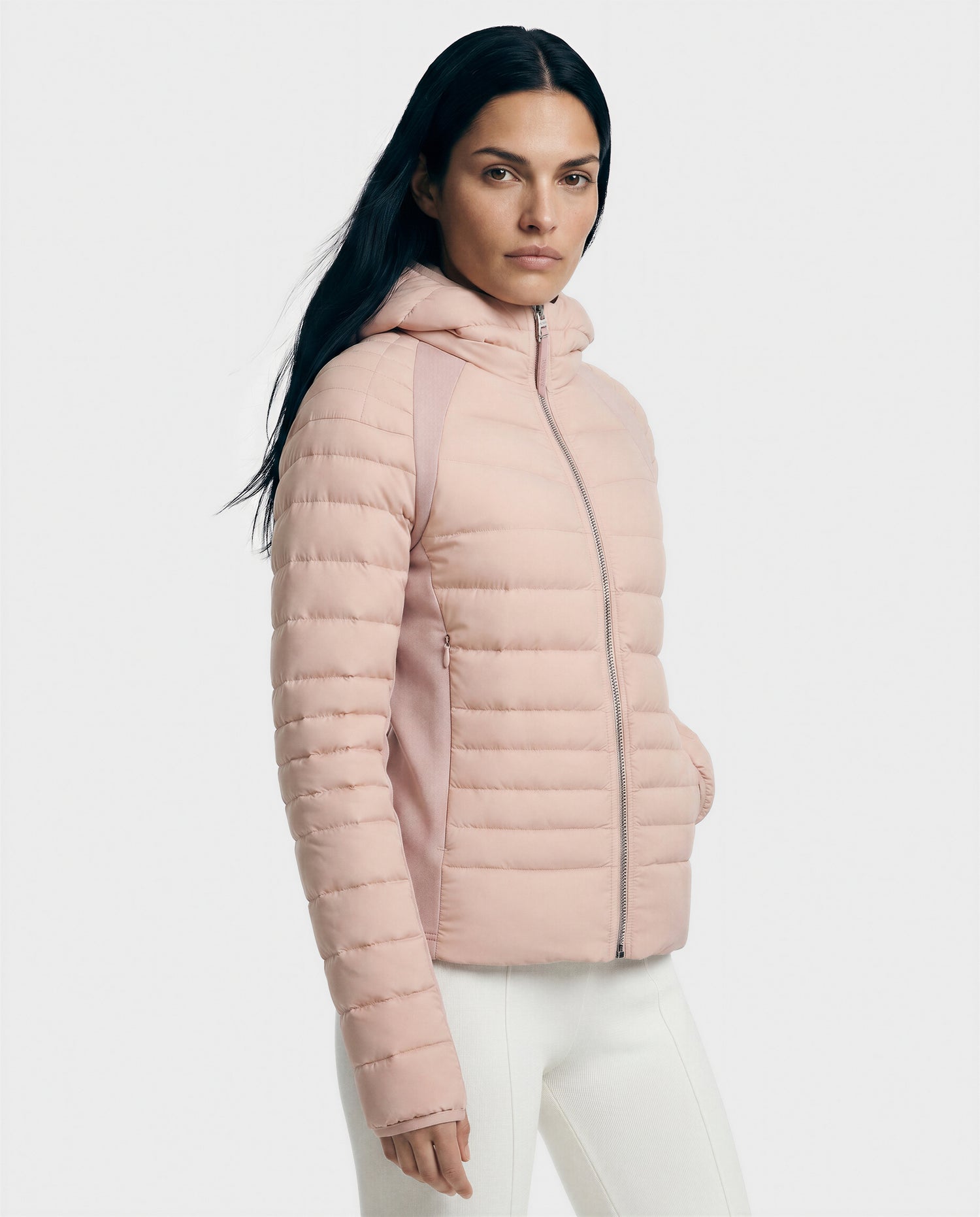 A woman with long dark hair models the Rudsak LENA - L124005 Soft Pink quilted jacket, featuring neoprene inserts and a hood, paired with white pants against a plain light background.
