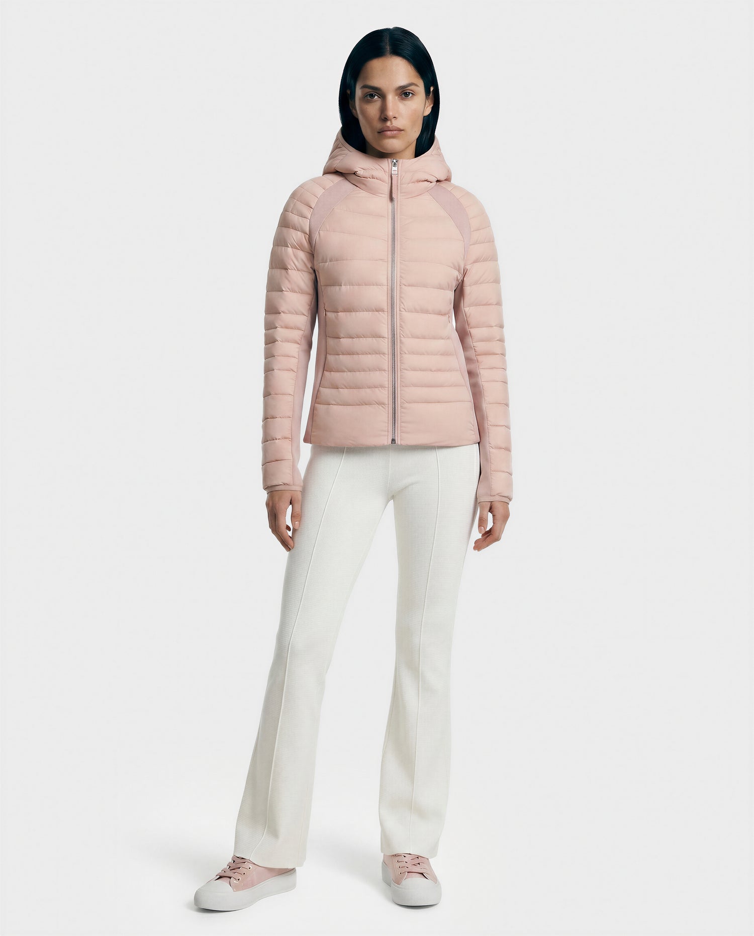 A woman faces forward, wearing the Rudsak LENA - L124005 Soft Pink quilted jacket with neoprene inserts, paired with white pants and pink sneakers, against a plain white background.