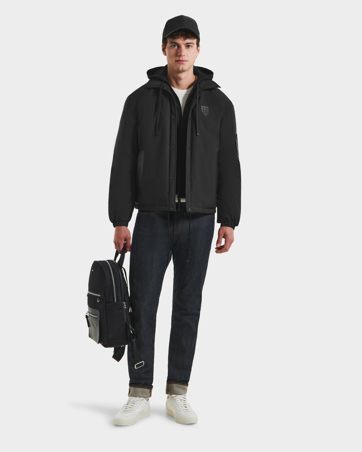 A young man in Rudsak’s LUCA HV - H125505 Black limited-edition jacket, dark jeans, white sneakers, and a black cap stands holding a black backpack, looking ahead against a plain white background.