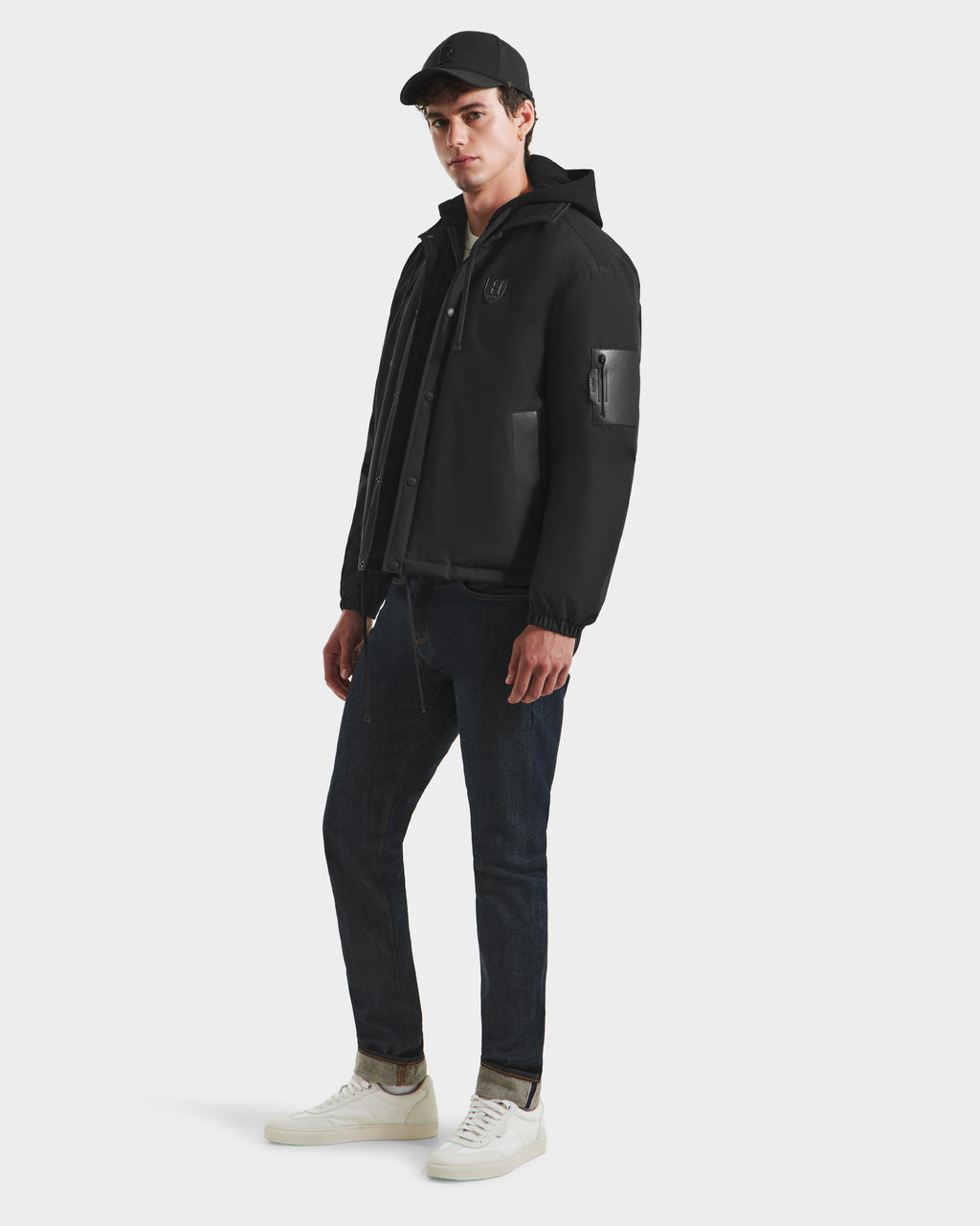 A man wearing the Rudsak LUCA HV - H125505 Black limited-edition hooded jacket, a black baseball cap, dark jeans, and white sneakers stands against a plain white background.