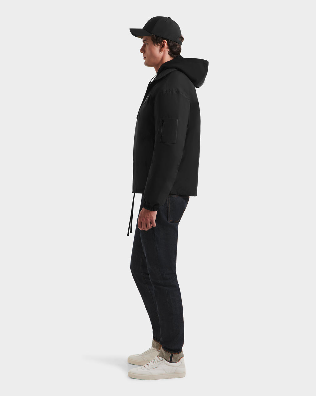 A man stands in profile facing right, wearing the Rudsak LUCA HV - H125505 Black hooded jacket, dark jeans, white sneakers, and a black baseball cap against a plain white background.