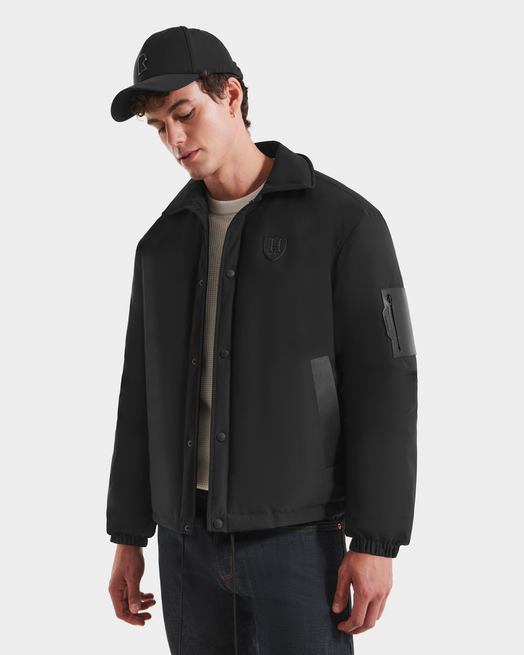 A young man wears the Rudsak LUCA HV - H125505 Black limited-edition jacket with a chest patch, paired with a black cap, beige sweater, and dark jeans. He stands against a white background, looking down with a relaxed expression.