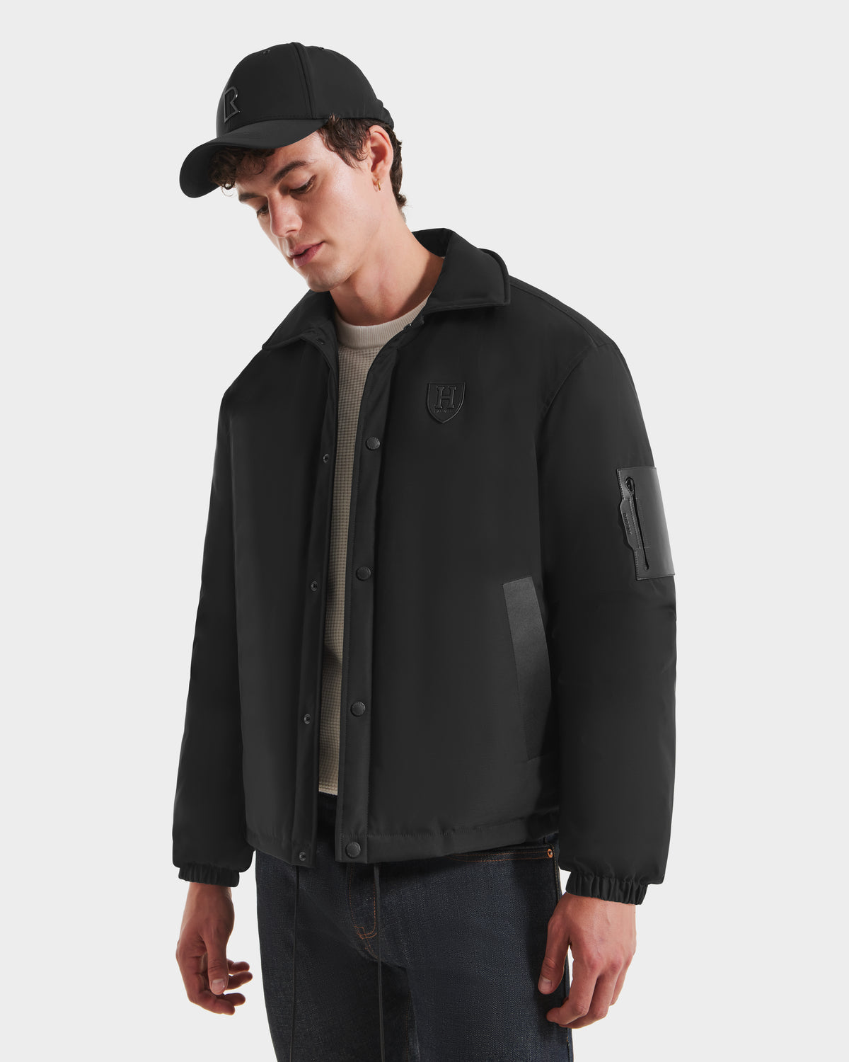 A young man wears the Rudsak LUCA HV - H125505 Black limited-edition jacket with a chest patch, paired with a black cap, beige sweater, and dark jeans. He stands against a white background, looking down with a relaxed expression.
