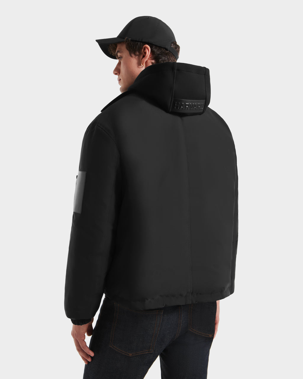A person in the Rudsak LUCA HV - H125505 Black limited-edition jacket with a raised collar featuring subtle text, dark jeans, and a black cap stands facing away from the camera against a plain light background.