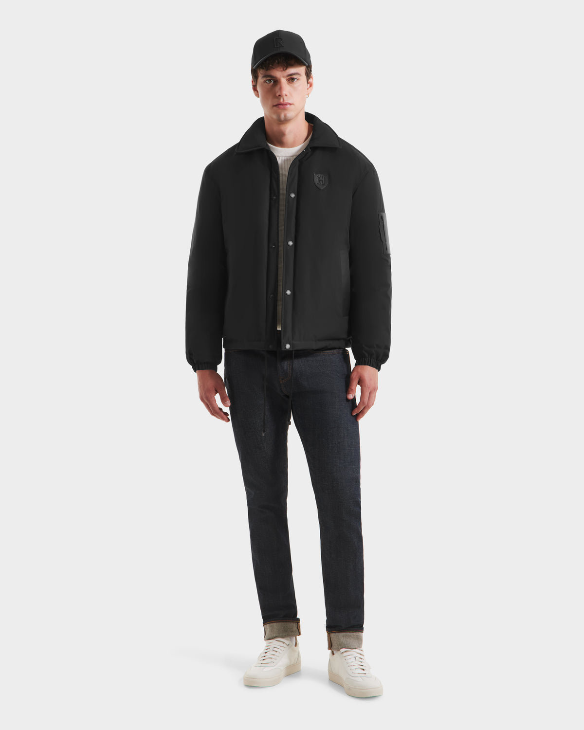 A young man stands facing forward against a white background, wearing the Rudsak LUCA HV - H125505 black jacket, a black cap, dark rolled-cuff jeans, white sneakers, and a white shirt.