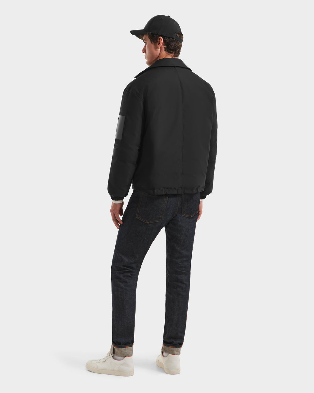 A man wearing the Rudsak LUCA HV - H125505 Black jacket, a black cap, dark jeans, and white sneakers stands facing away, looking to the left against a plain white background.