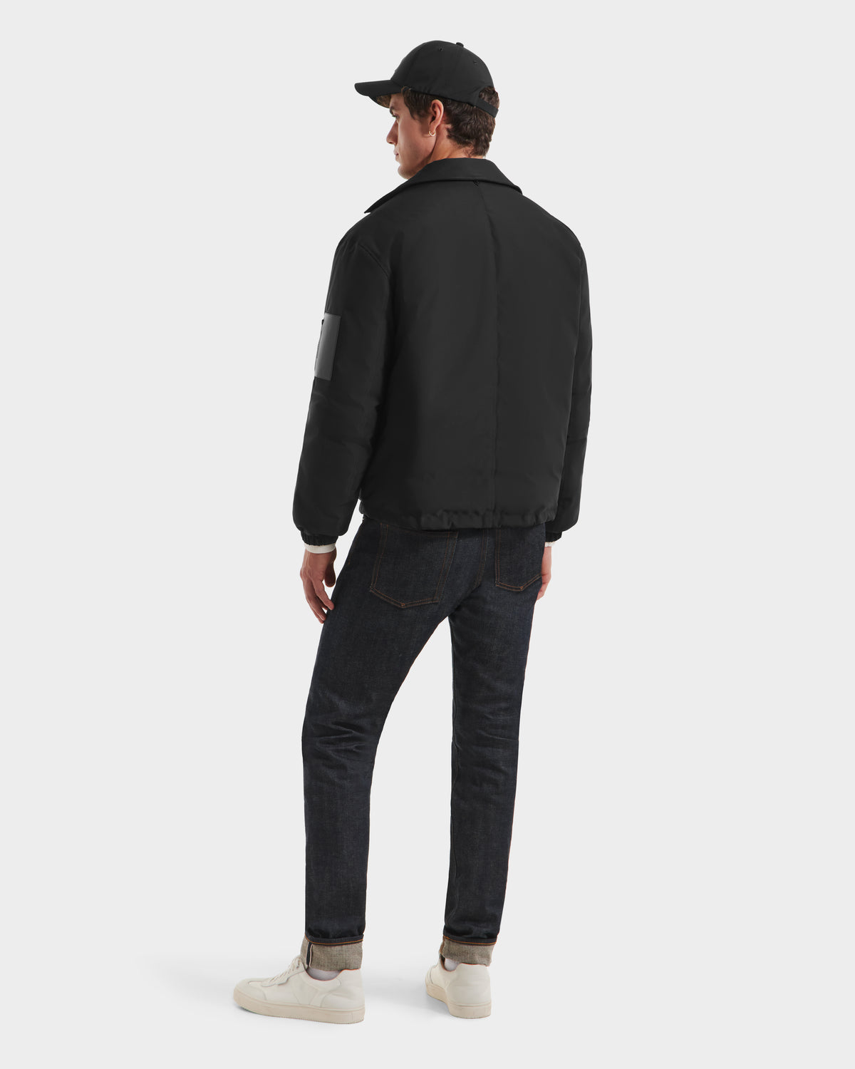 A man wearing the Rudsak LUCA HV - H125505 Black jacket, a black cap, dark jeans, and white sneakers stands facing away, looking to the left against a plain white background.