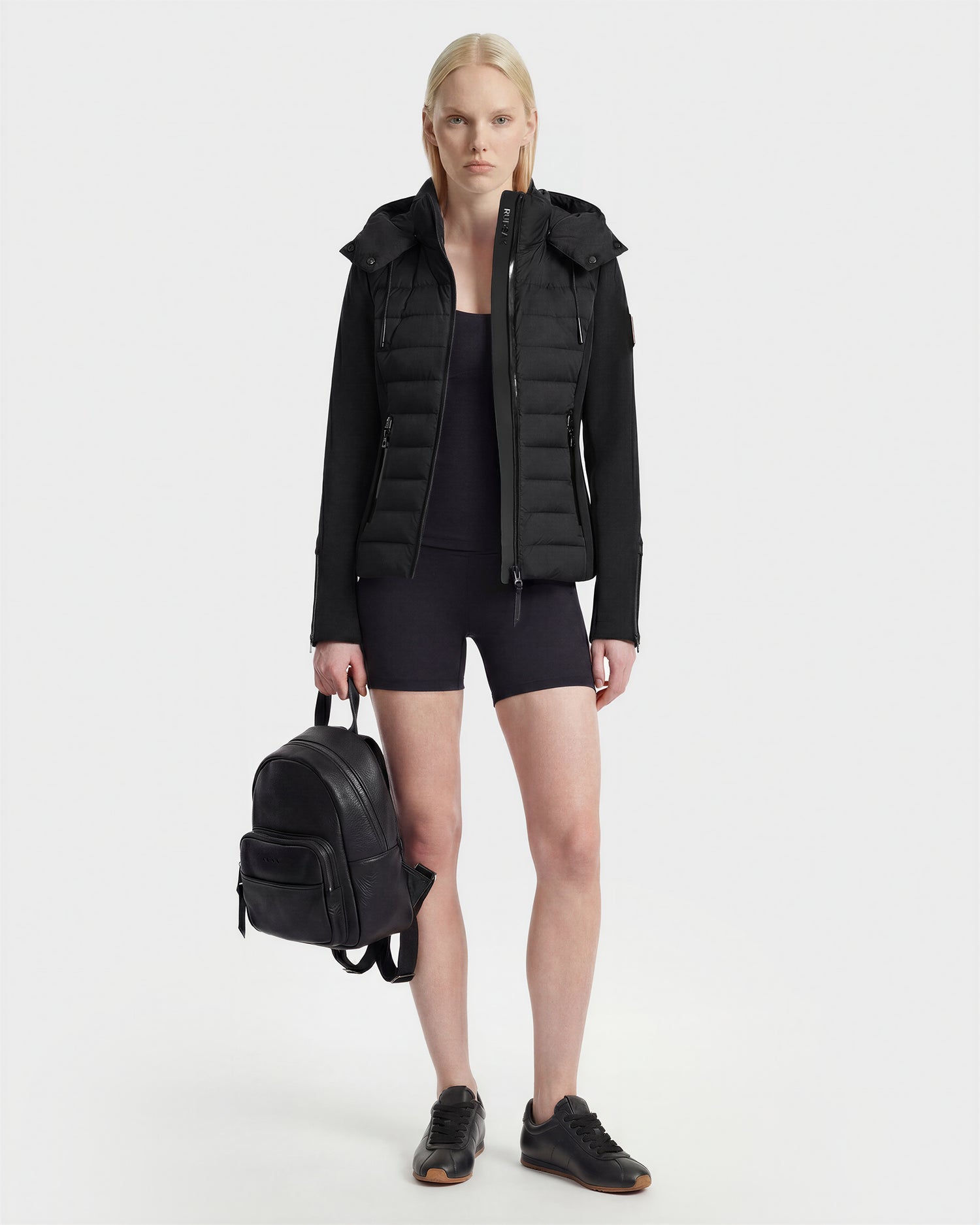 A woman with long blonde hair models the Rudsak MABS - 8125055 Black puffer jacket over a black top and cycling shorts, holding a black backpack and wearing black sneakers against a plain white background.