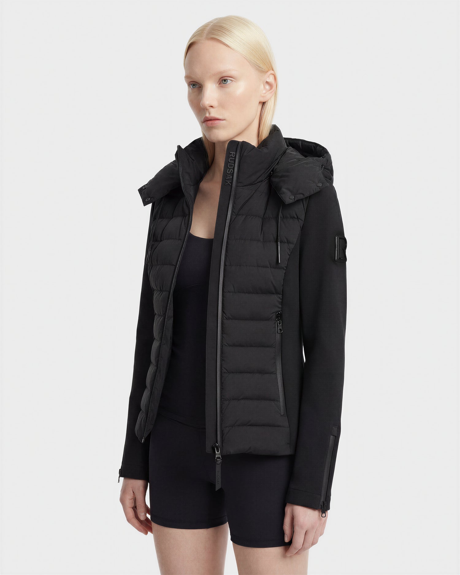 A woman with straight blonde hair wears the Rudsak MABS - 8125055 Black women’s puffer jacket over a black top and shorts, standing against a plain light background.