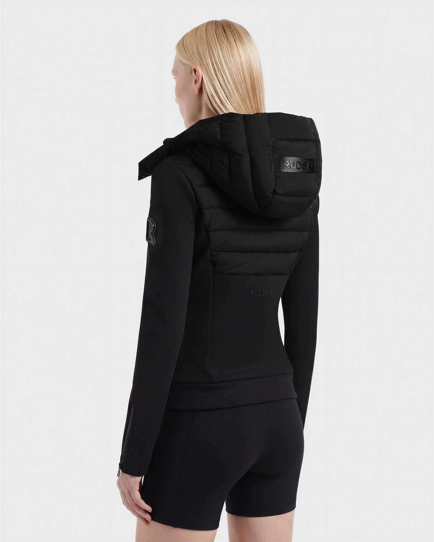 A person with straight blonde hair is shown from behind against a plain white background, wearing Rudsak’s MABS - 8125055 Black lightweight activewear jacket and black shorts.