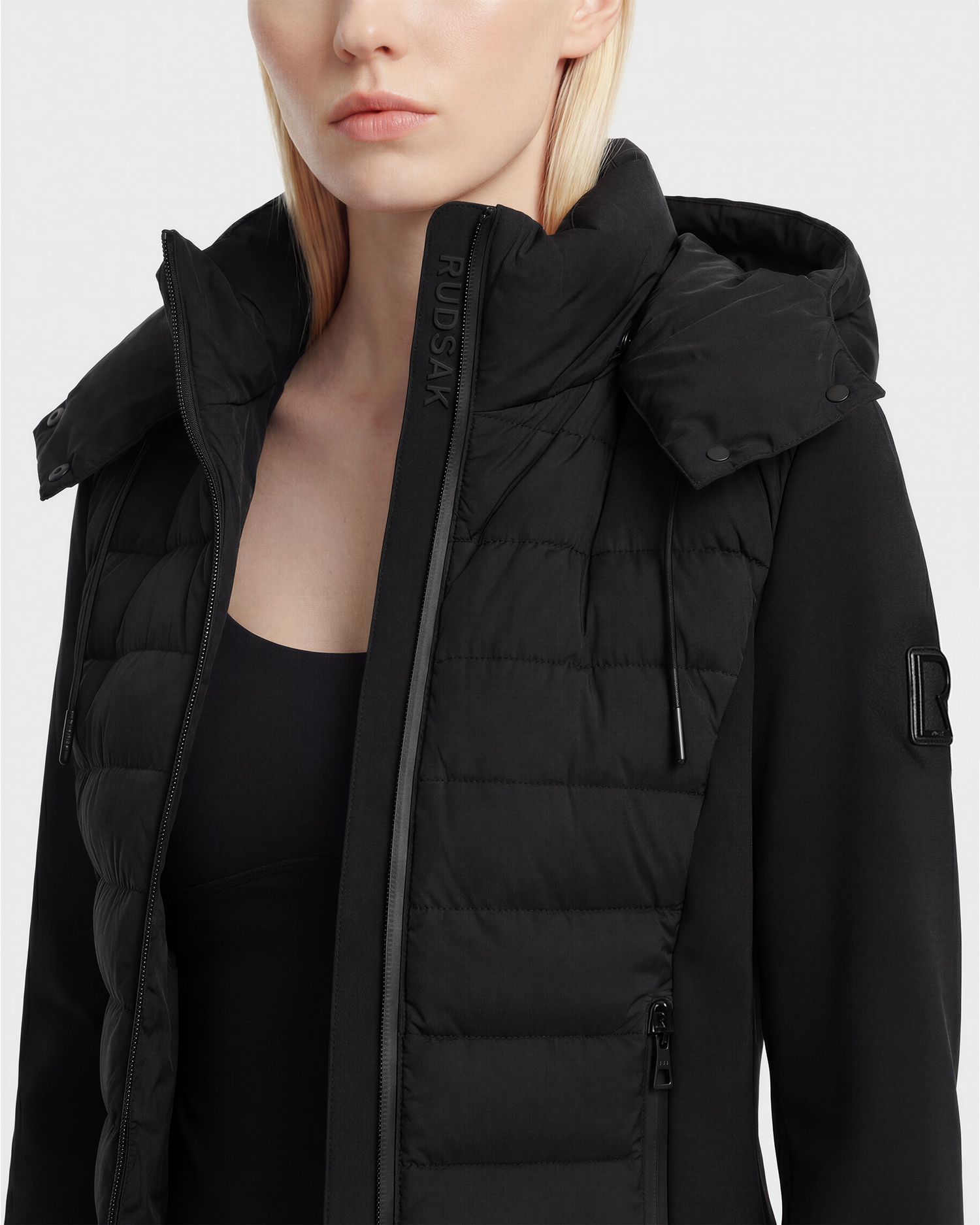 A woman with straight blonde hair wears the Rudsak MABS - 8125055 Black women’s puffer jacket—RDS-certified down, sleeve patch visible—over a black top against a plain, light background.