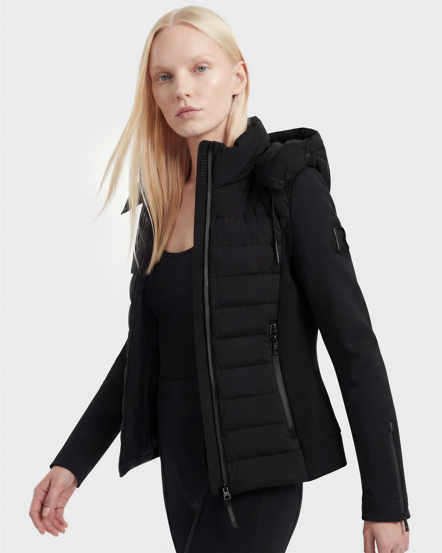 A woman with long, straight blonde hair models the Rudsak MABS - 8125055 Black lightweight activewear jacket with zippered pockets over a black top and pants, posing against a plain light gray background.