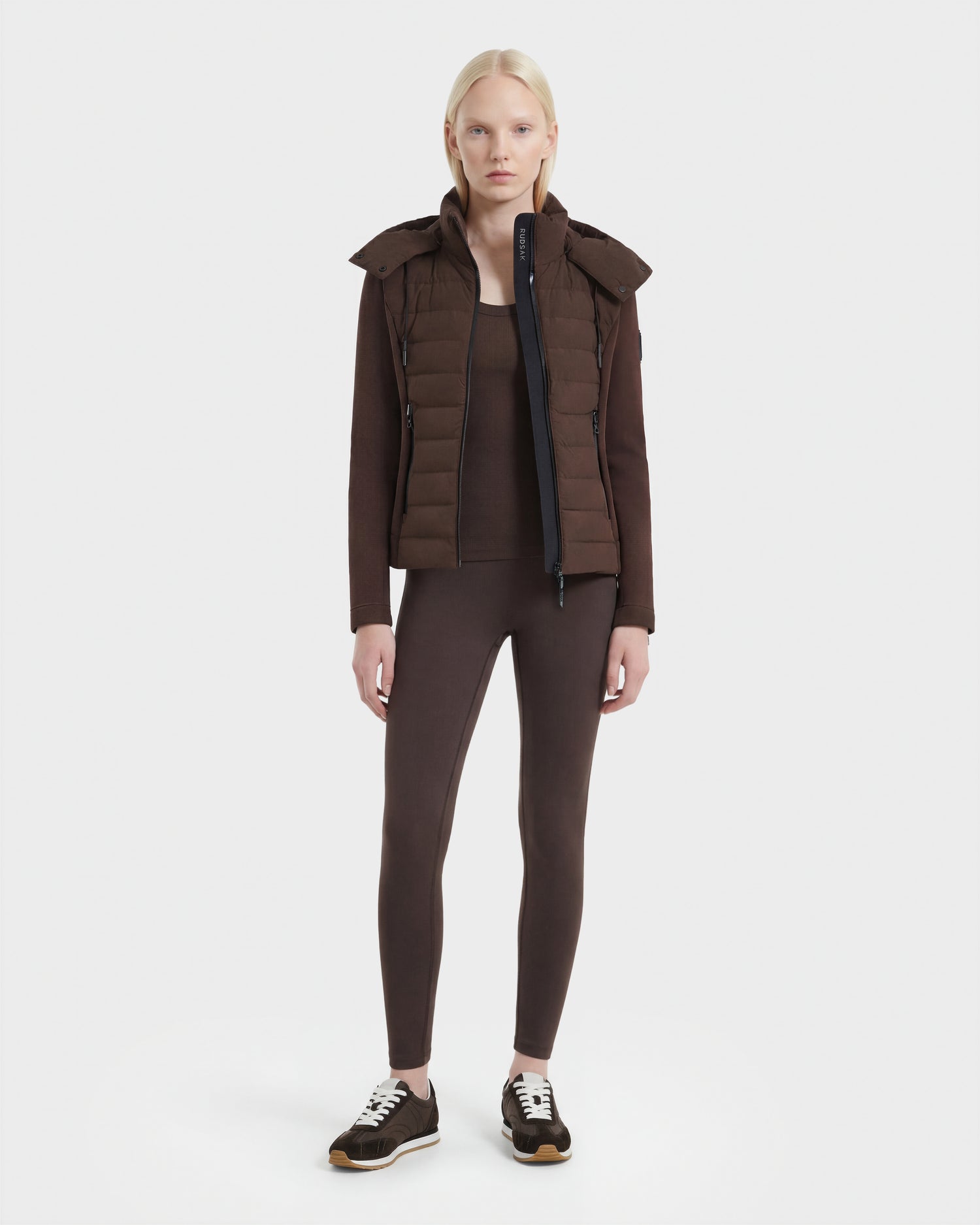 A woman with straight blonde hair faces forward, modeling the Rudsak MABS - 8124825 Espresso: a lightweight short jacket over matching brown long-sleeve top and leggings, paired with black sneakers, against a plain white background.