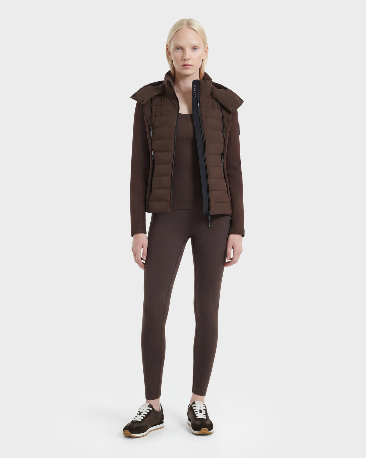 A woman with straight blonde hair faces forward, modeling the Rudsak MABS - 8124825 Espresso: a lightweight short jacket over matching brown long-sleeve top and leggings, paired with black sneakers, against a plain white background.