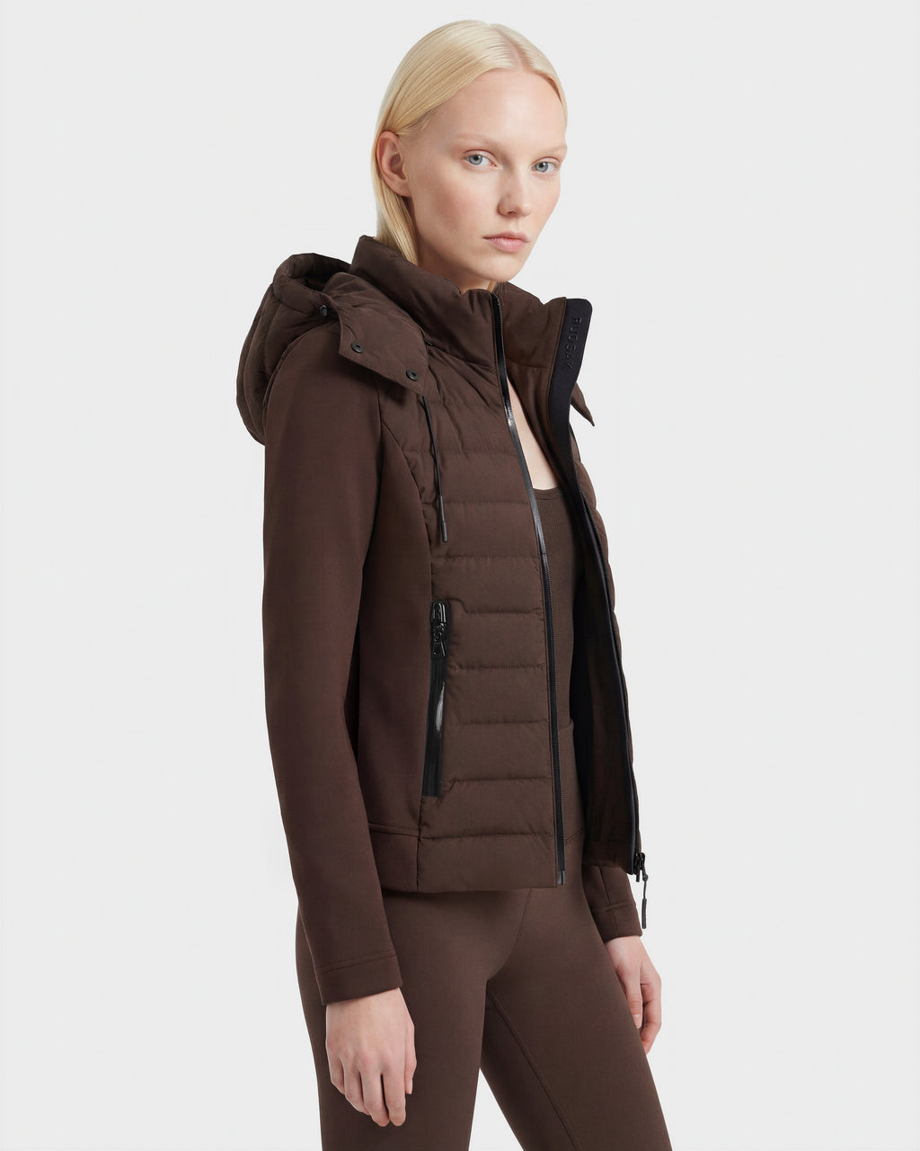 A woman with straight blonde hair wears the Rudsak MABS - 8124825 Espresso hooded puffer jacket and matching pants, standing against a plain white background and facing slightly to the side.