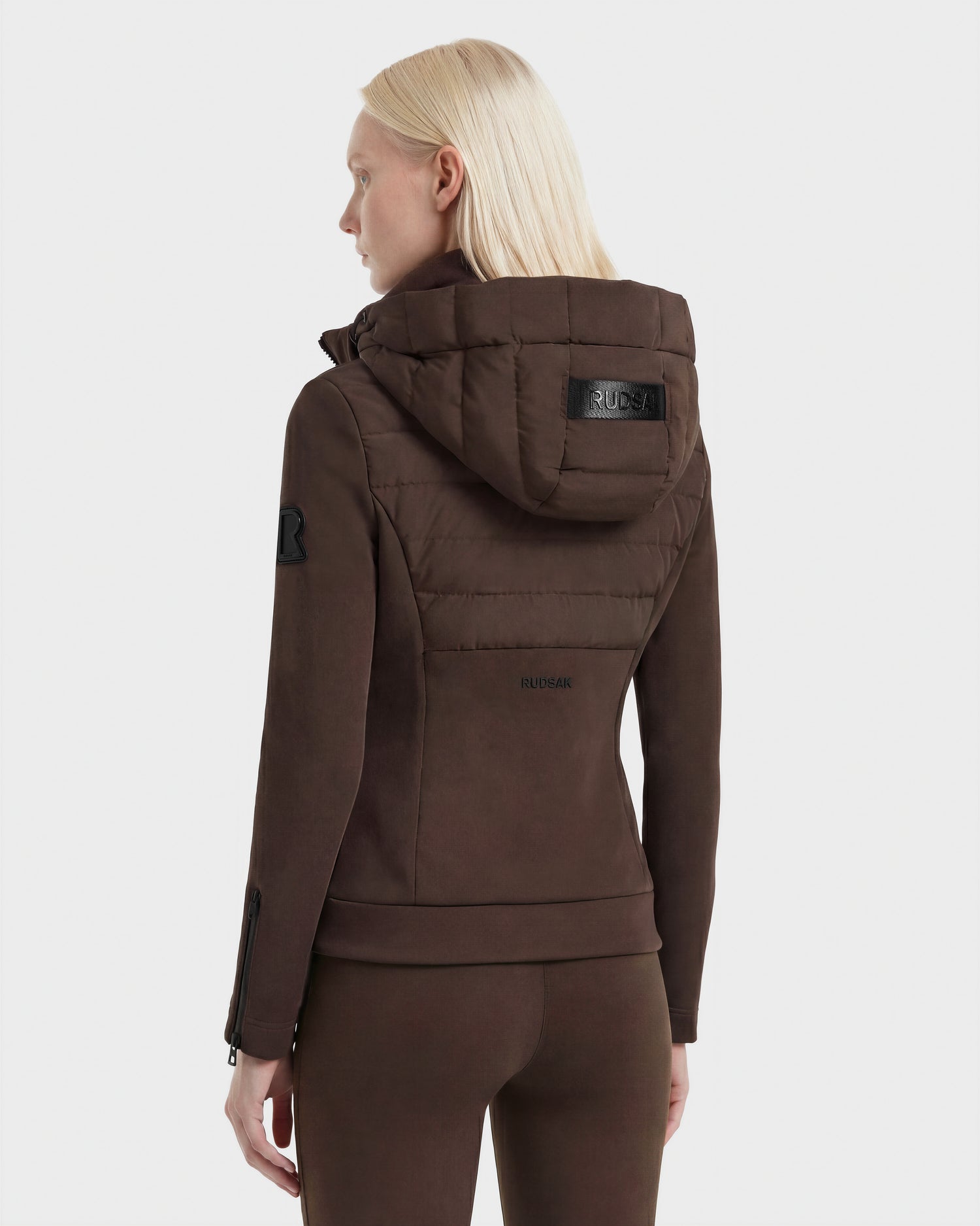 A woman with straight blonde hair is shown from the back, wearing the Rudsak MABS - 8124825 Espresso lightweight short jacket and matching pants. The neoprene jacket features a shiny black hood patch and small upper arm logo against a plain white background.
