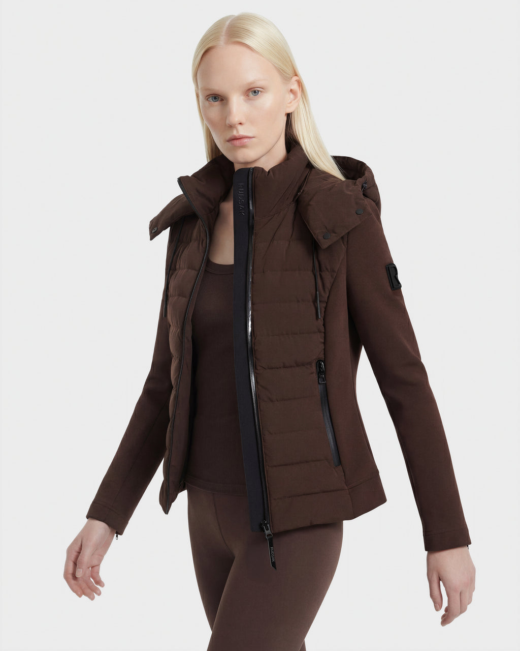 A woman with long blonde hair models the Rudsak MABS - 8124825 Espresso: a lightweight short jacket with detachable hood, styled over a dark brown top and matching pants, against a plain white background.