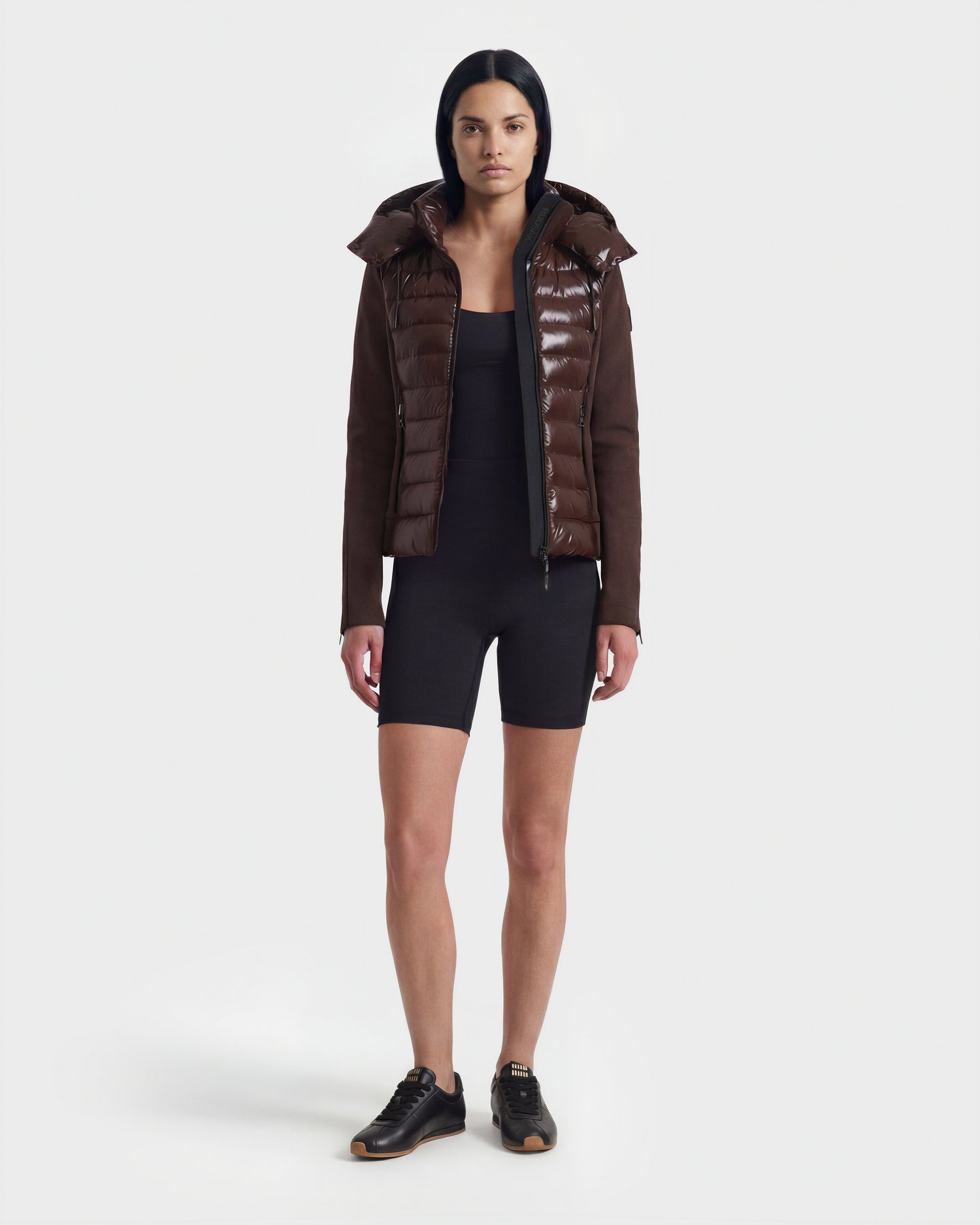 A woman stands facing forward against a plain white background wearing the Rudsak MABS C - 8124590 Espresso puffer over a black fitted romper and black sneakers, her arms relaxed by her sides.