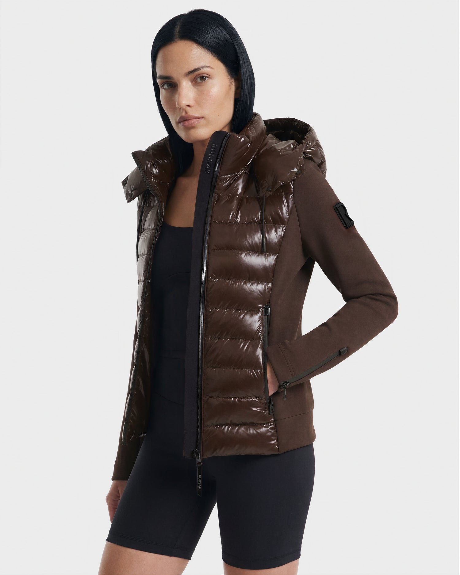 A woman with straight black hair wears the Rudsak MABS C - 8124590 Espresso shiny brown women’s puffer jacket over a fitted black outfit, standing against a plain light background.