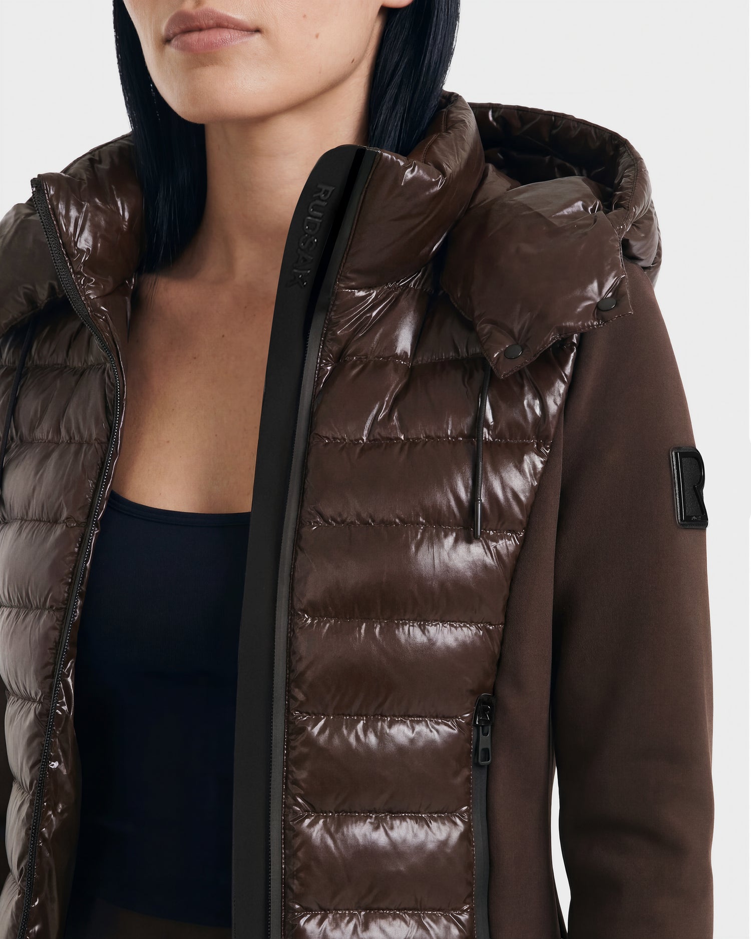 A woman stands against a light background wearing the Rudsak MABS C - 8124590 Espresso puffer jacket, a brown shiny hooded zip-up with a fabric sleeve patch and black top underneath.