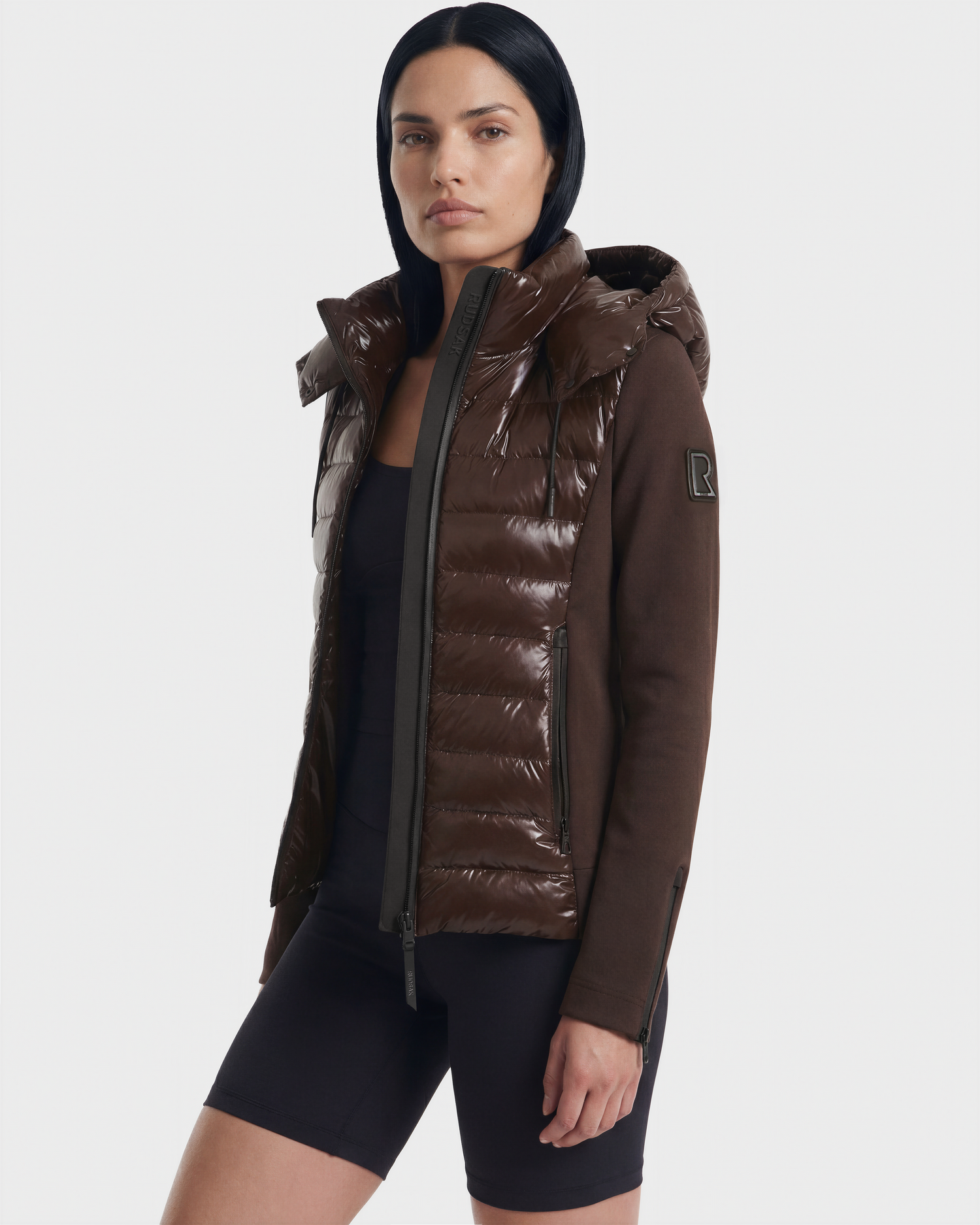 A woman with straight dark hair wears the Rudsak MABS C - 8124590 Espresso shiny brown puffer jacket with a hood over a black top and shorts, standing against a plain white background.