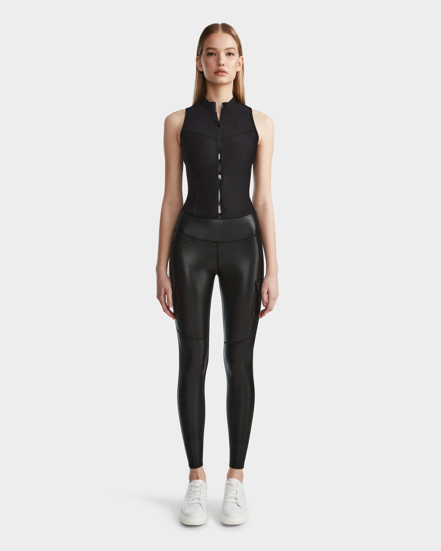 A woman faces forward against a light background, wearing the Rudsak SIEN L - 8524079 Black sleeveless zip-up top and black high-waisted vegan leather leggings, paired with white sneakers.