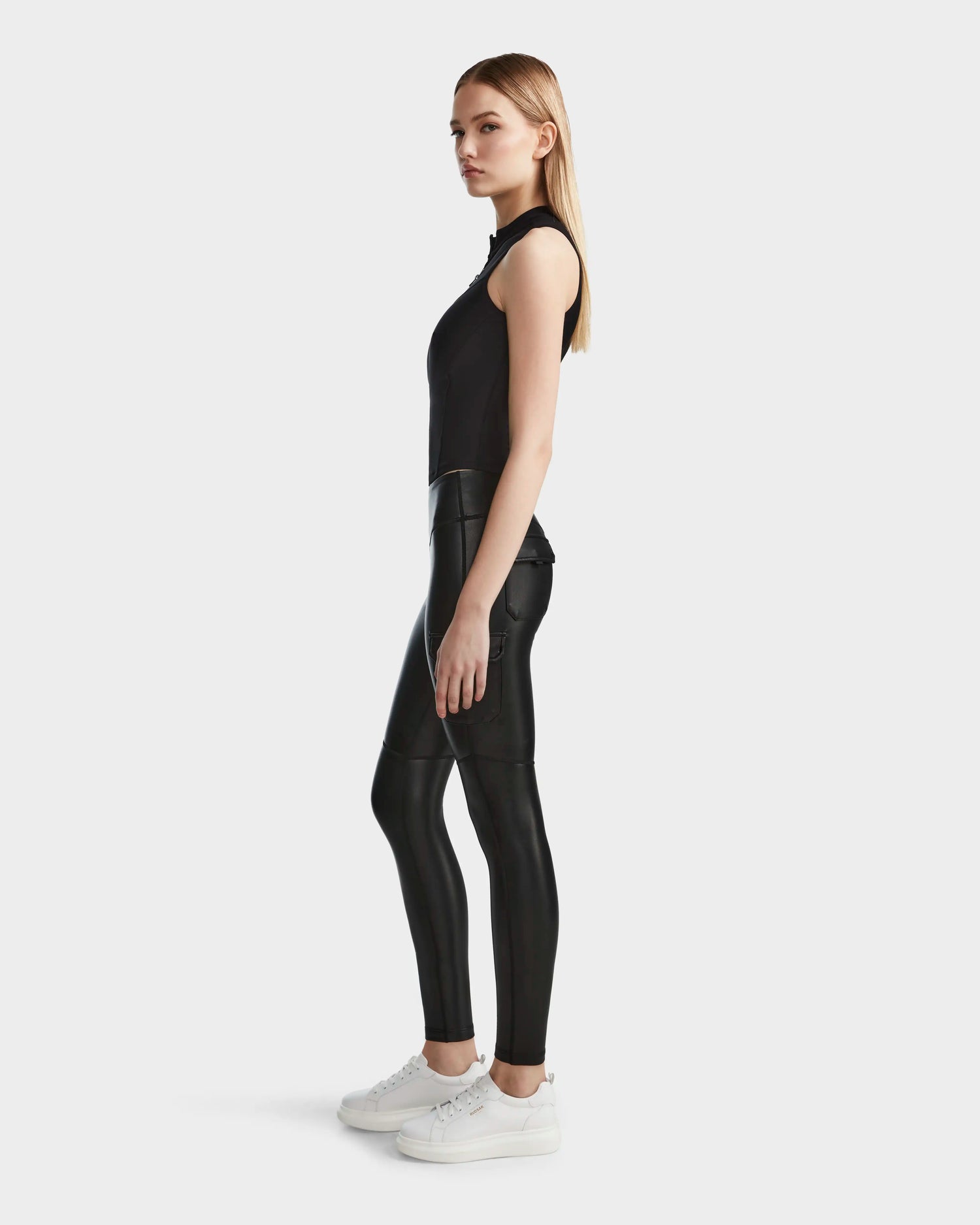 A woman with straight long blonde hair and a neutral expression stands in profile against a plain background, wearing Rudsak’s SIEN L - 8524079 Black sleeveless top, shiny vegan leather leggings, and white sneakers.