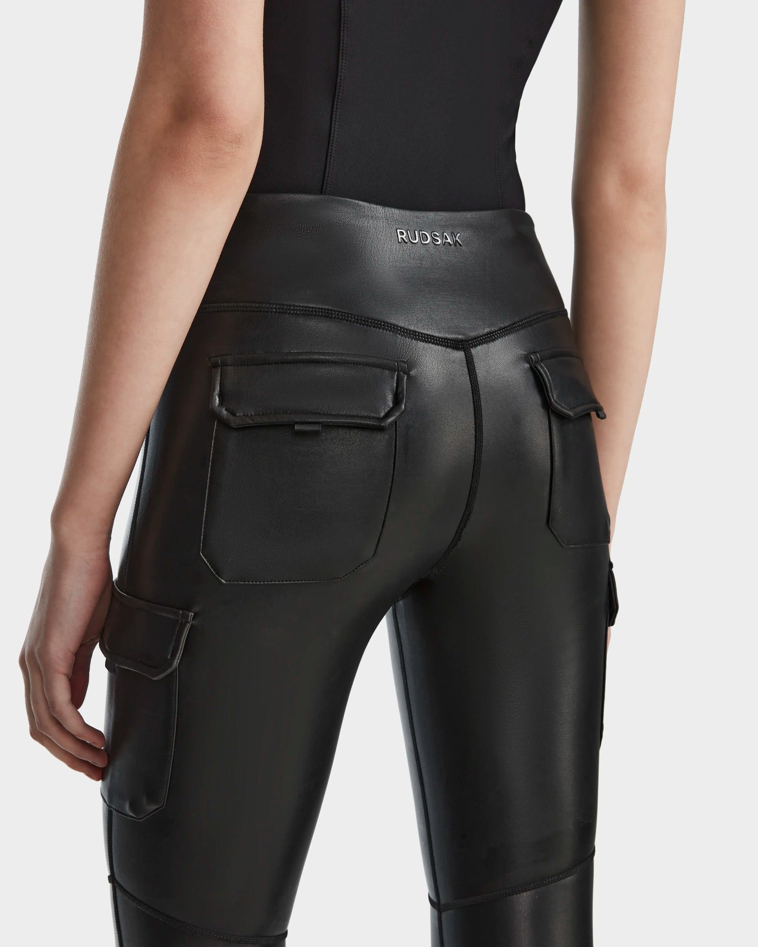 A person is seen from the back wearing Rudsak SIEN L - 8524079 black cargo leggings with a shiny vegan leather finish, large pockets, and a fitted waistband featuring the Rudsak logo for an edgy activewear look.