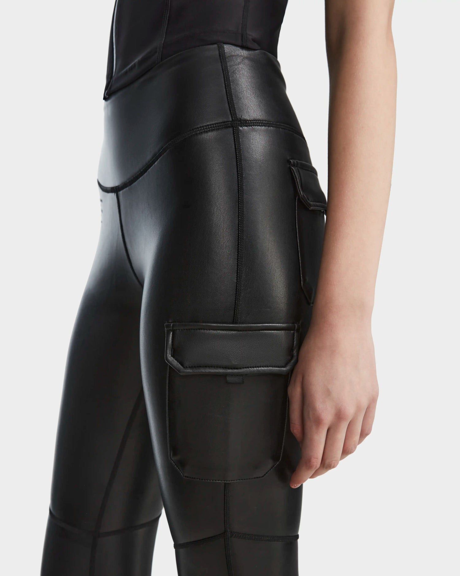 A person wearing Rudsak SIEN L - 8524079 Black high-waisted vegan leather cargo leggings with large side pockets, shown waist down with their right arm relaxed at their side.