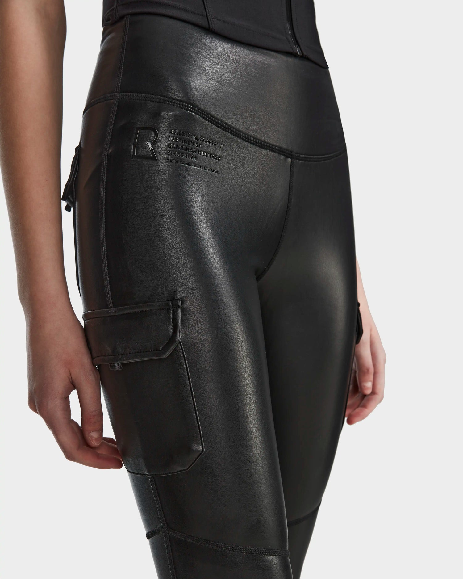 A person wears Rudsak's SIEN L - 8524079 black high-waisted stretch vegan leather leggings with a side pocket and embossed hip detail, shown waist to mid-thigh against a plain background—ideal for elevating women’s activewear style.