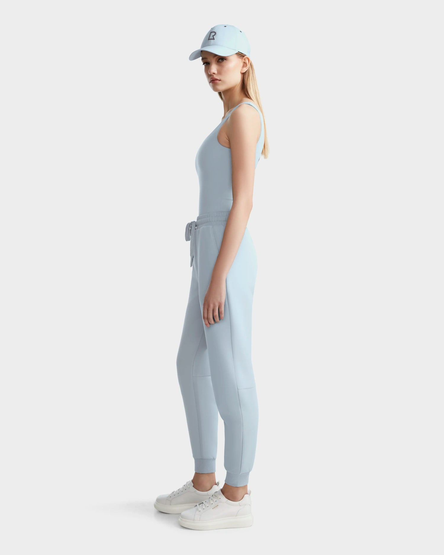 A woman stands sideways against a plain white background, looking at the camera and wearing Rudsak’s BRIDGET - 8524033 Sky Blue tank bodysuit, jogger pants, a matching cap, and white sneakers.