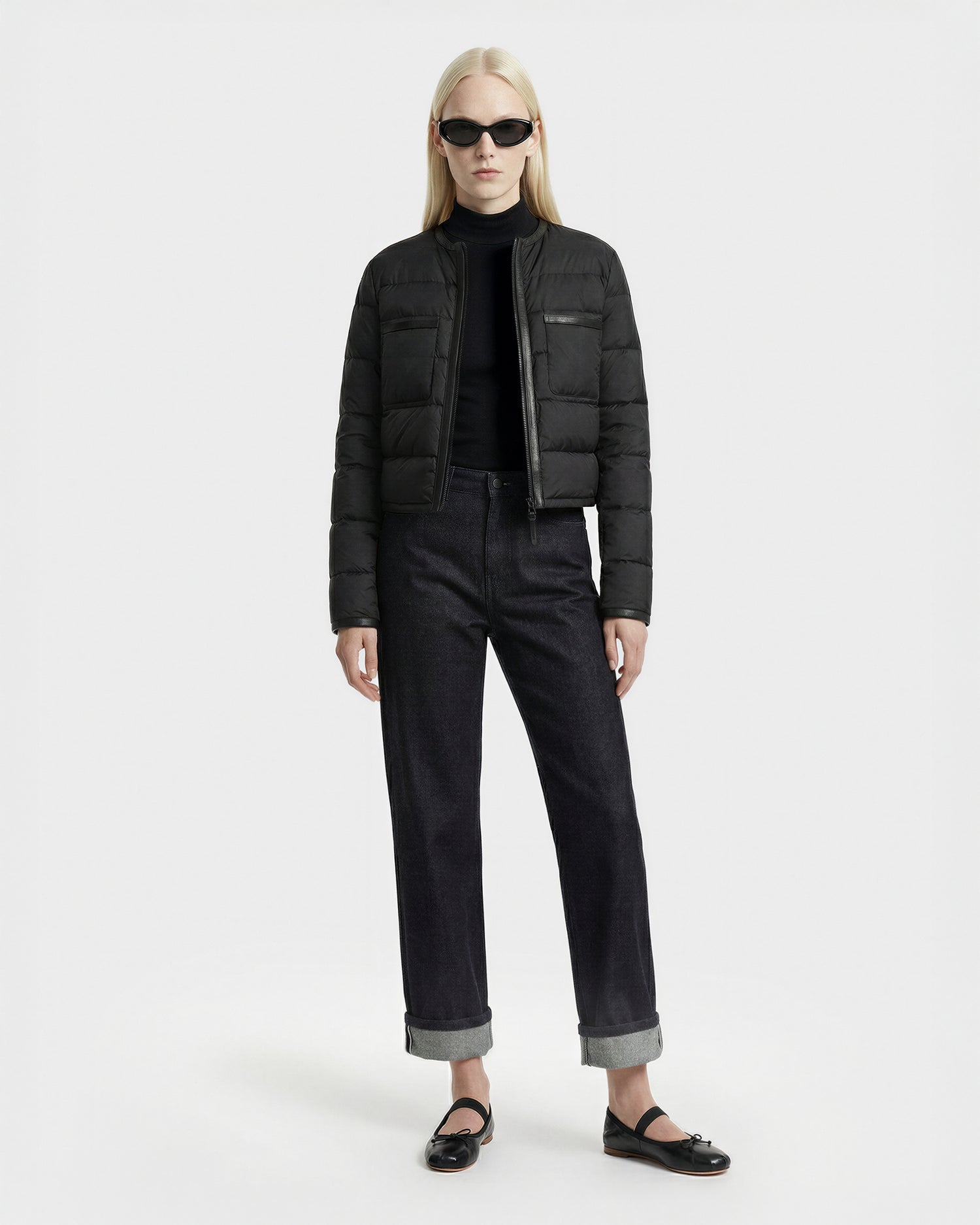 A person with long blonde hair wears black sunglasses and the Rudsak MARKLE - 8125033 Black women’s cropped down jacket, paired with a black turtleneck, dark cuffed jeans, and black flats against a plain white background.