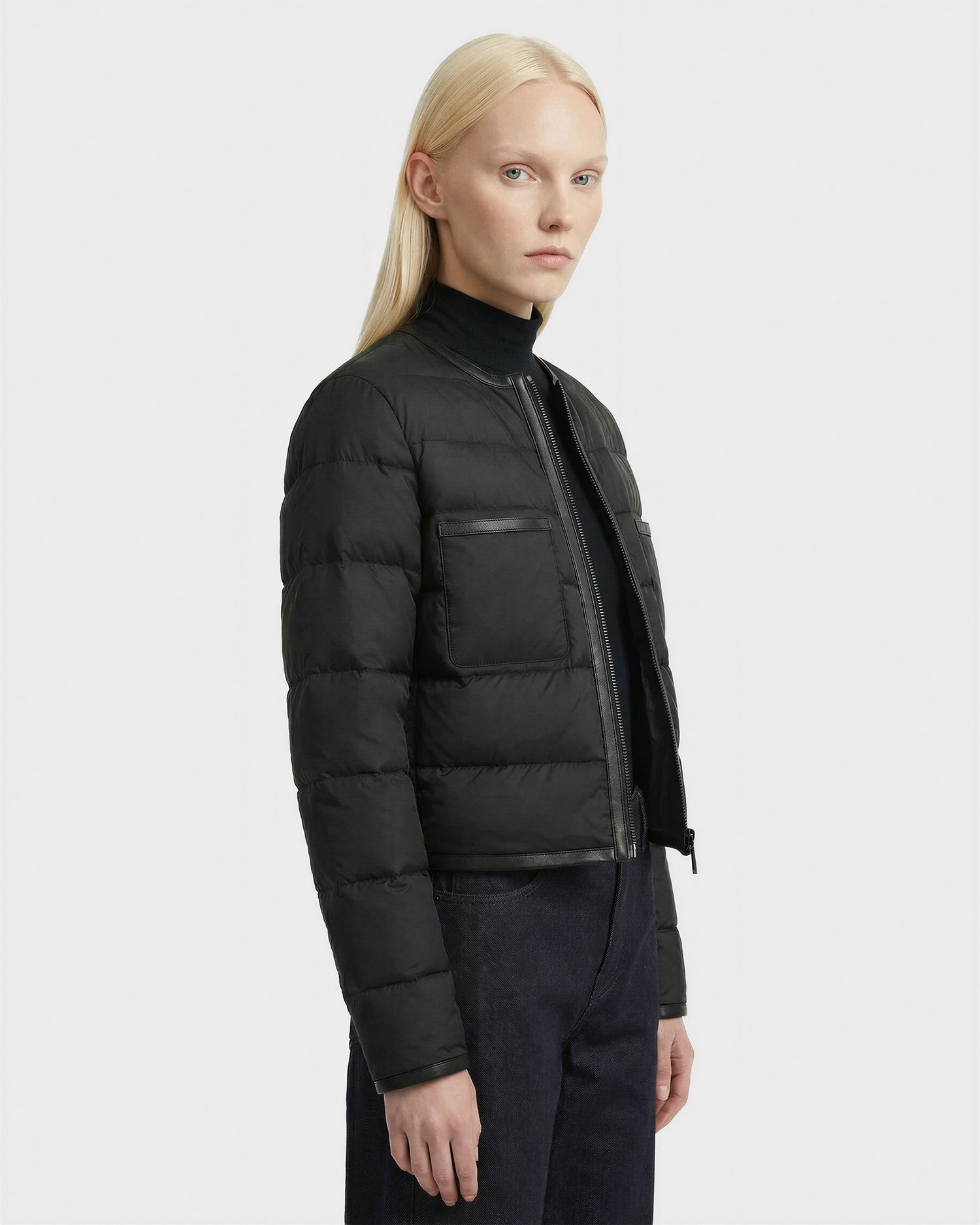 A person with long blond hair wears the Rudsak MARKLE - 8125033 Black women’s cropped down jacket with vegan leather trim, a black turtleneck, and dark pants, standing sideways against a plain white background.