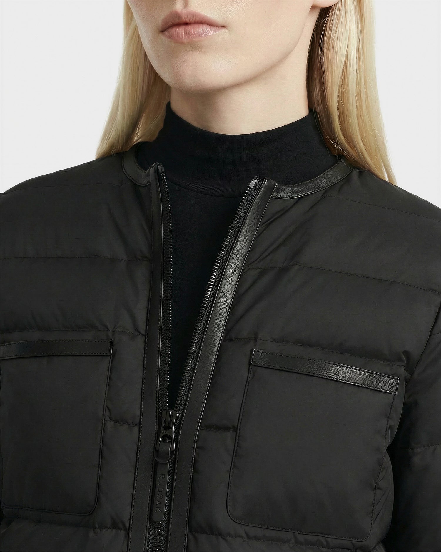 A woman with straight blonde hair models the Rudsak MARKLE - 8125033 Black lightweight down jacket, featuring a front zipper and two chest pockets, over a black turtleneck. The photo is cropped just below her nose.