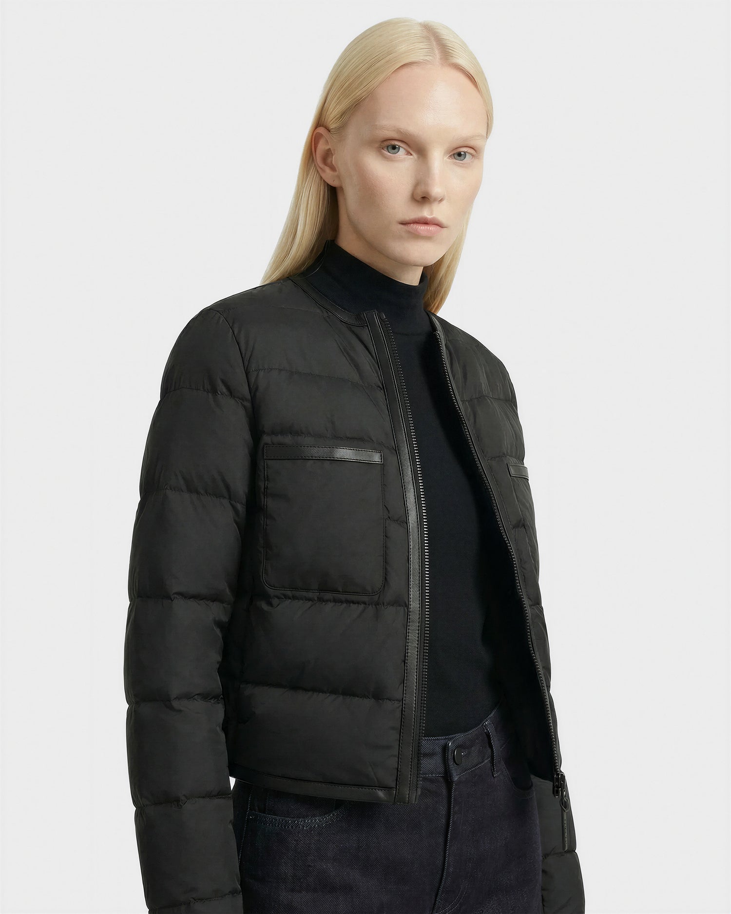 A person with straight, long blonde hair wears the Rudsak MARKLE - 8125033 Black down jacket over a black turtleneck and dark jeans, standing against a plain light gray background.
