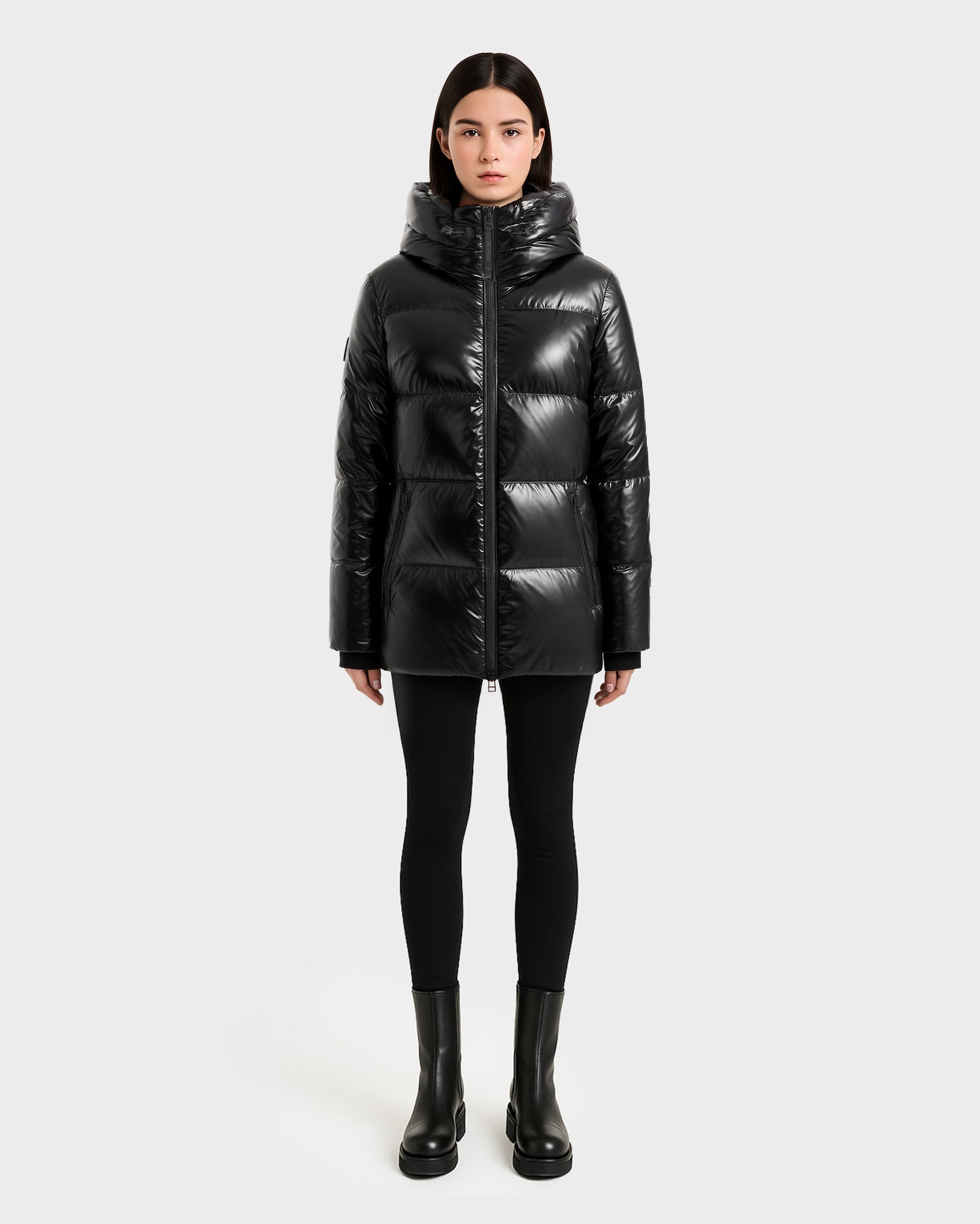 A woman faces forward in the Rudsak MARY - 4124500 Black puffer jacket with a high collar and hood, styled with black leggings and ankle boots against a plain, light background.