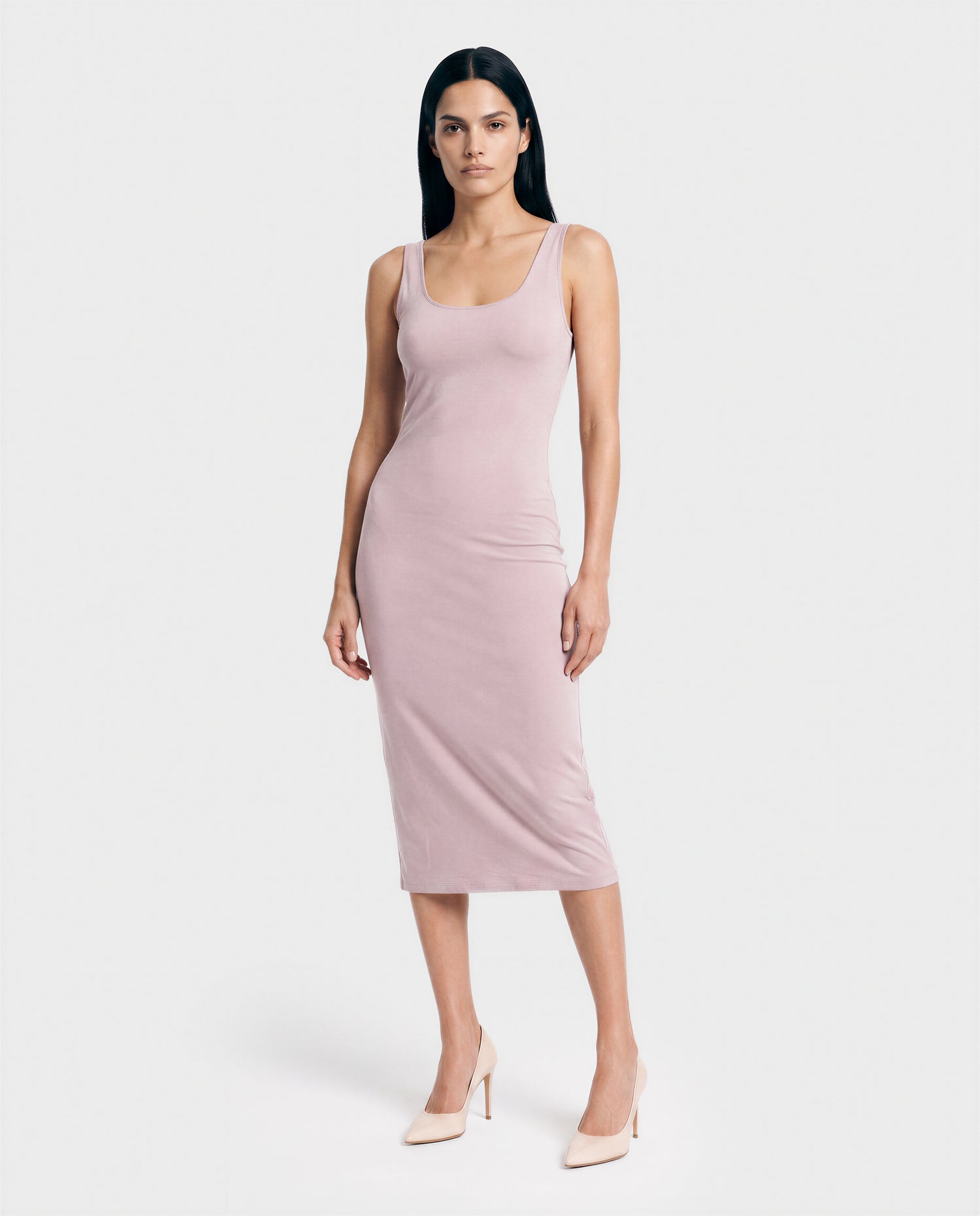 A woman with straight dark hair stands against a white background, wearing the Rudsak AIMEE - L523025 dress in soft pink and nude heels, her arms relaxed by her sides and a neutral expression on her face.