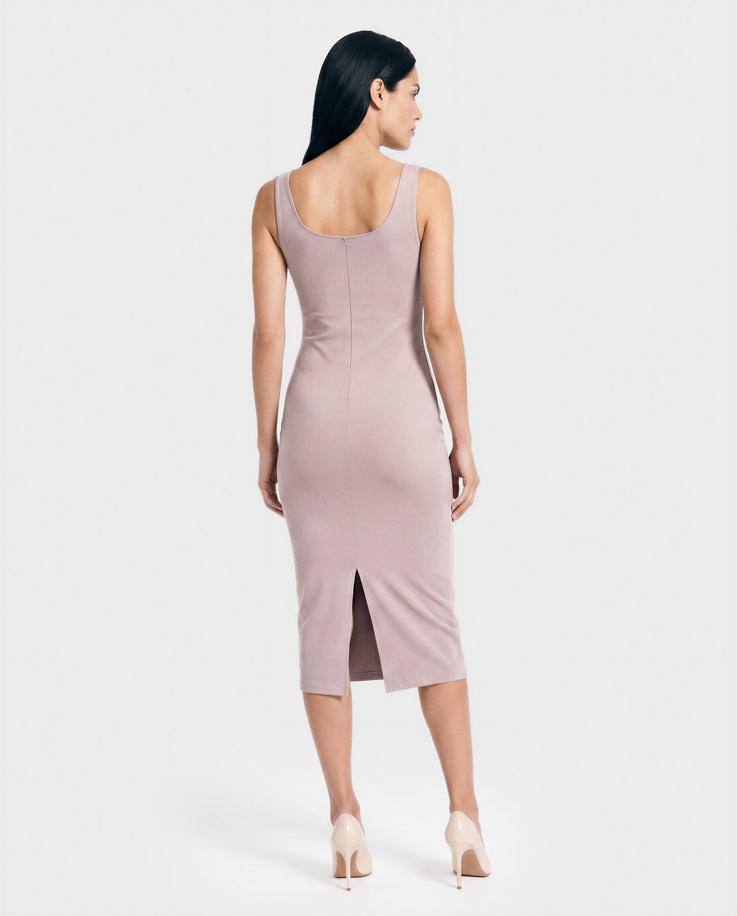 A woman with long dark hair faces away, wearing the AIMEE - L523025 SOFT PINK mid-length stretch dress by Rudsak, featuring a back slit and nude heels, crafted from premium stretch fabric against a plain light background.