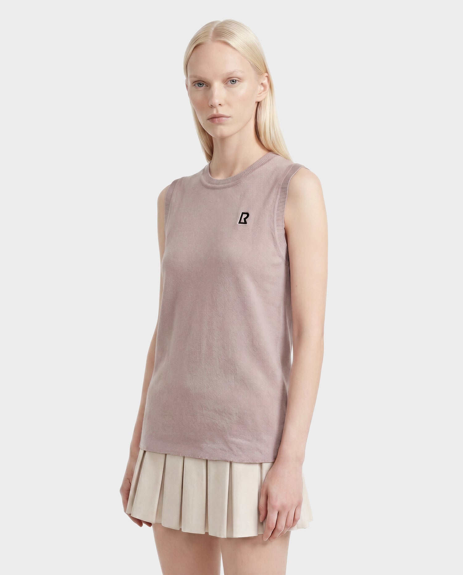 A woman with long blonde hair wears the Rudsak LISA - L523024 minimalist sleeveless knit top in soft pink with a small black logo on the chest and a white pleated skirt, standing against a plain light background.