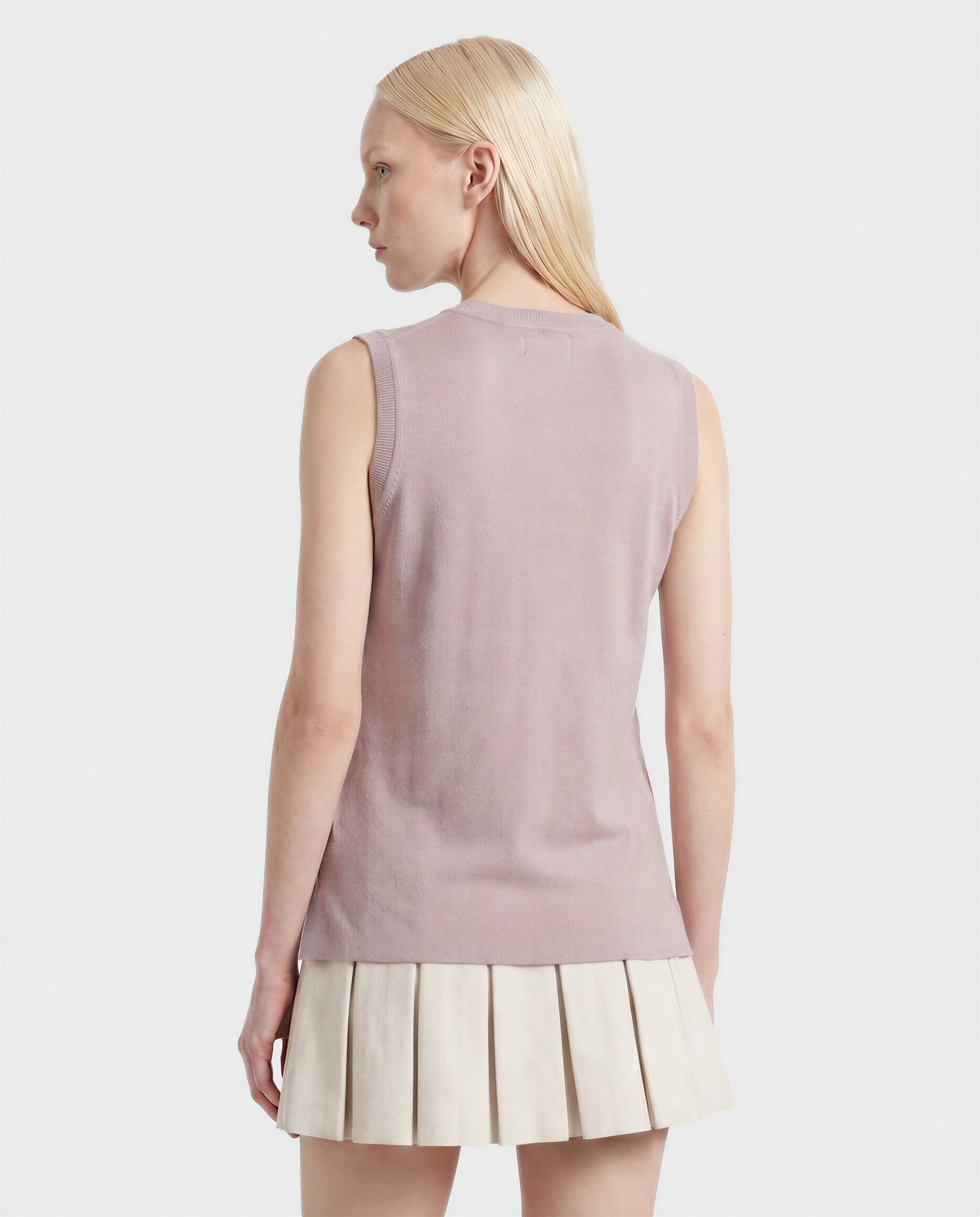 A person with long, straight blonde hair is seen from behind wearing the Rudsak LISA - L523024 SOFT PINK sleeveless knit top and a cream pleated skirt, standing against a plain white background.