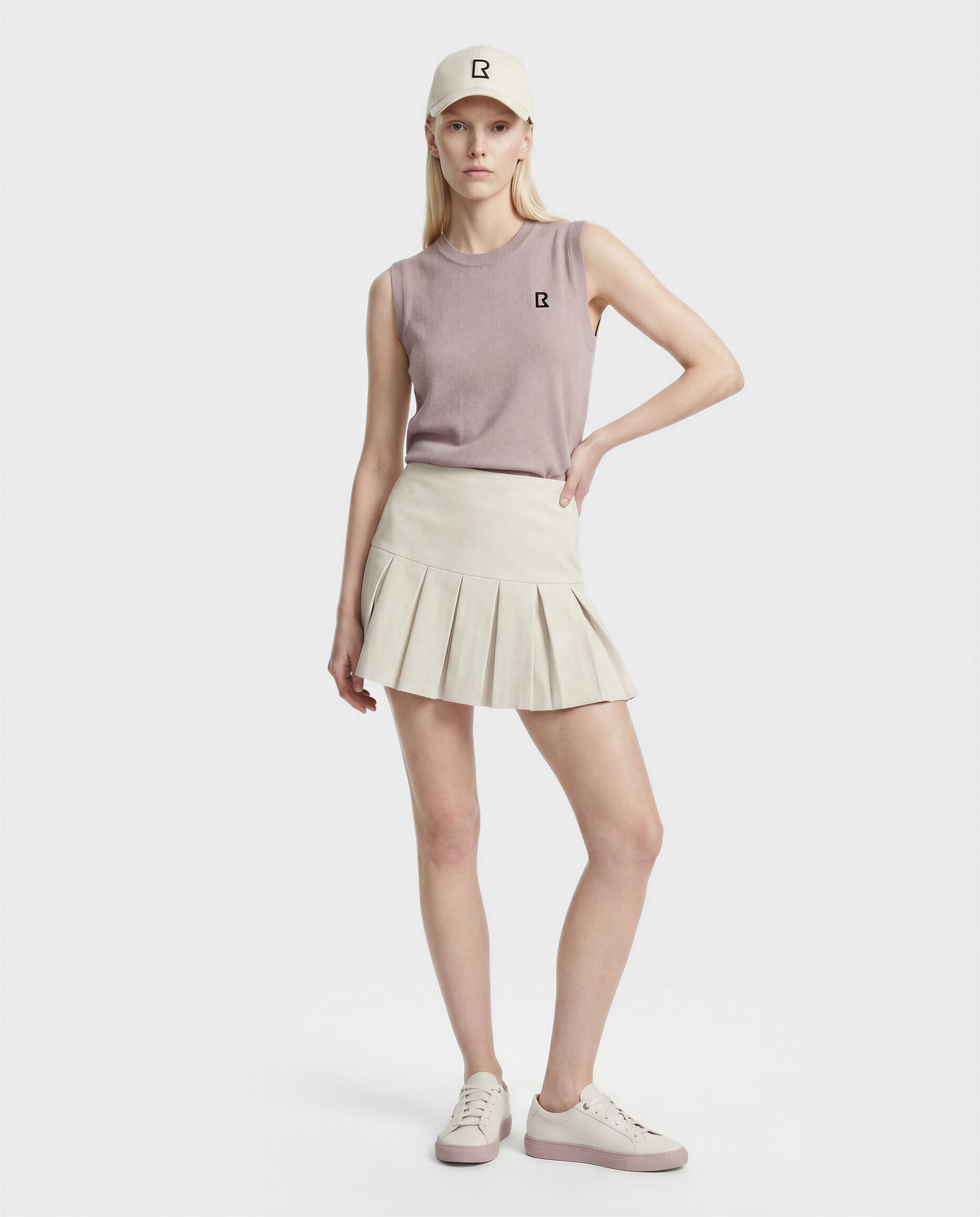 A woman with long blonde hair and a neutral expression wears Rudsak's LISA - L523024 SOFT PINK pleated skirt, a mauve sleeveless knit top, matching sneakers, and a beige cap against a white background, with one hand on her hip.
