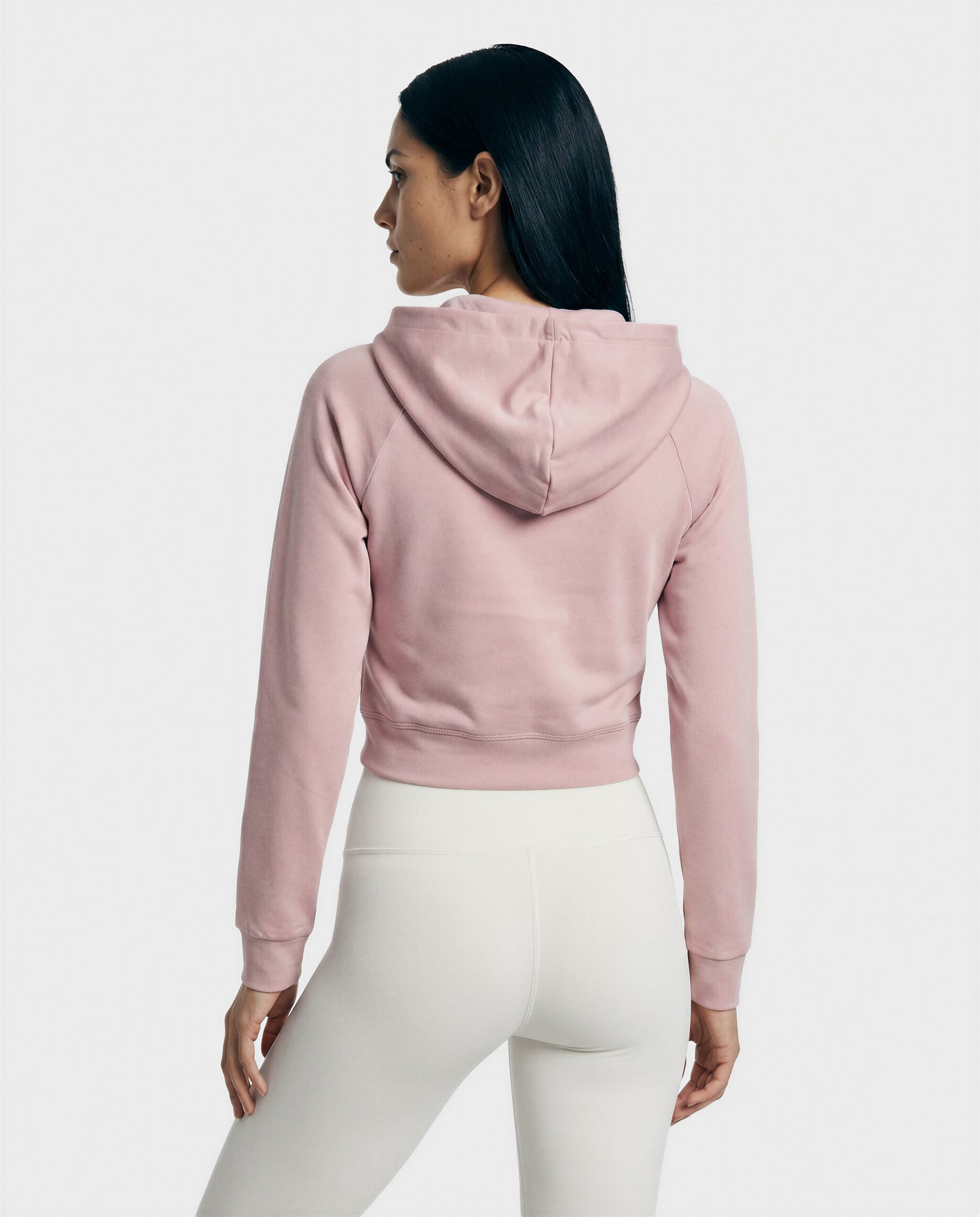 A woman with long dark hair faces away, wearing the Rudsak SAMMY - L523014T top in soft pink and high-waisted white leggings—a modern, effortless choice against a plain light background.