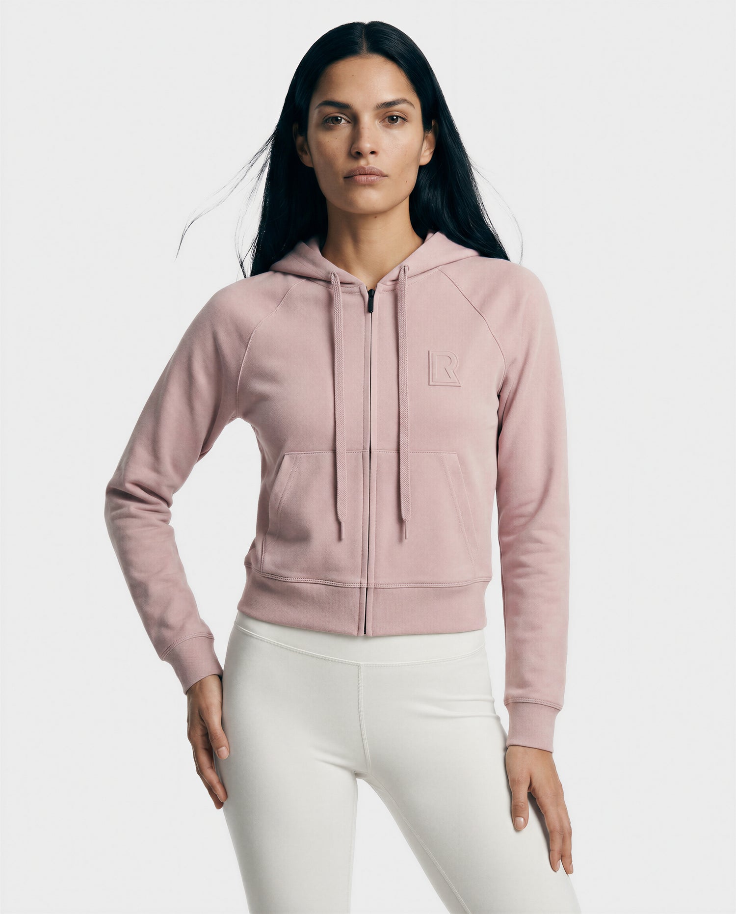 A woman with long dark hair wears the Rudsak SAMMY - L523014T soft pink zip-up crop top with a subtle chest logo and white fitted pants, standing against a plain white background—a stylish pick for modern everyday wear.