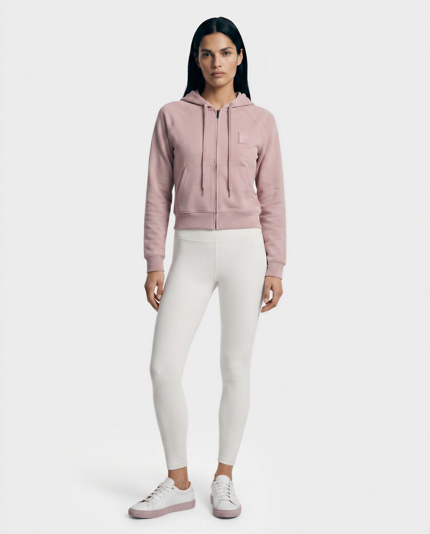 A woman models the Rudsak SAMMY - L523014T top in soft pink, paired with white leggings and white sneakers with pink soles, creating a fresh, modern everyday look against a plain light background.