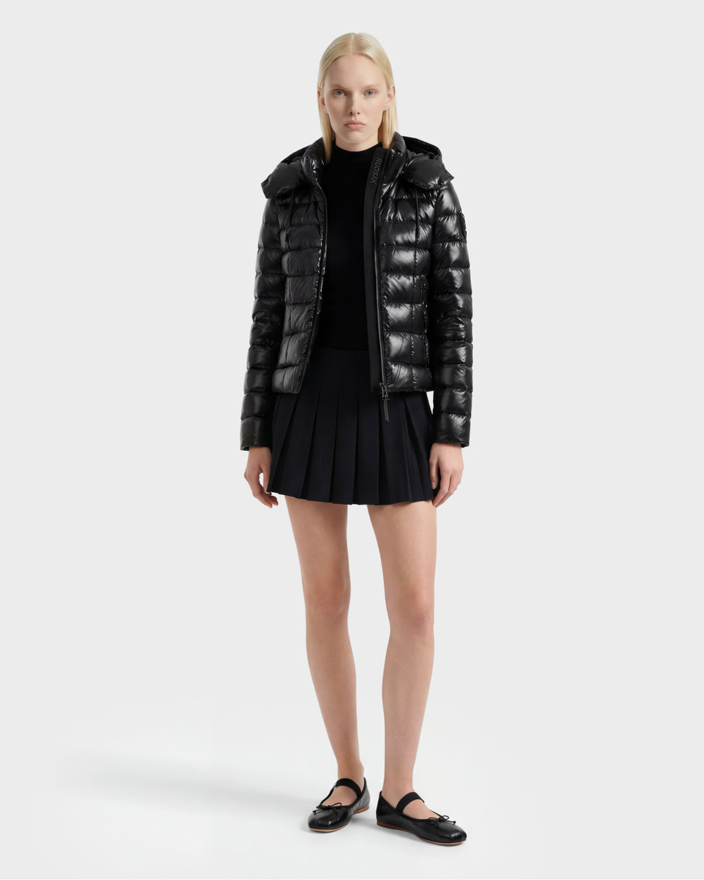 A woman with straight blonde hair and a neutral expression stands facing forward against a plain white background, wearing the Rudsak MELISSA C - 8125695 Black lightweight down puffer, a black top, pleated mini skirt, and strap flat shoes.