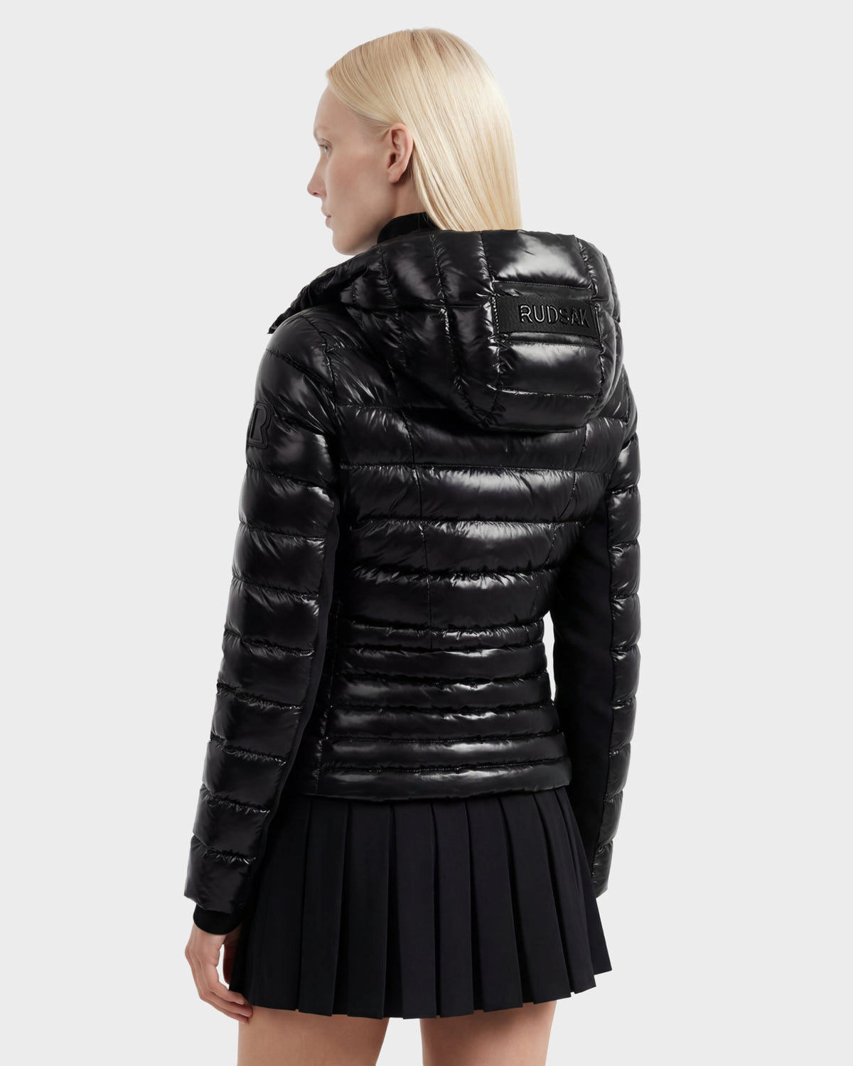 A person with straight, light blonde hair wears the MELISSA C - 8125695 Black women’s puffer jacket by Rudsak, featuring a hood and back branding, paired with a black pleated skirt against a plain light background.